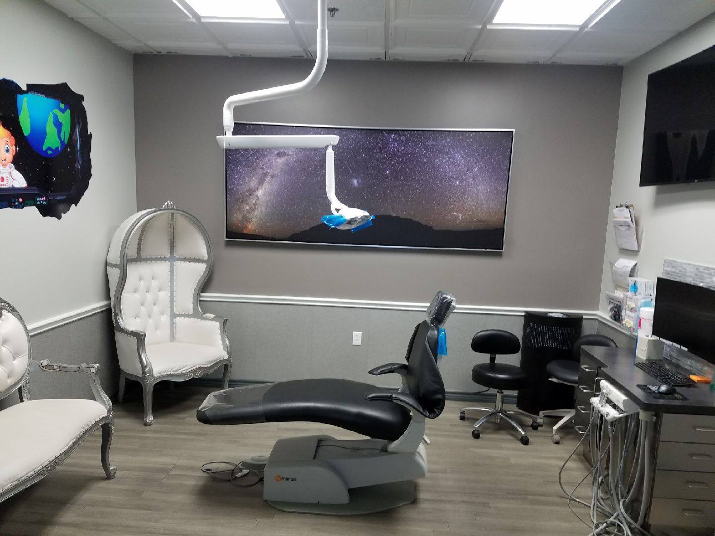 Dental exam room with chair, overhead monitor, and medical equipment on gray walls.