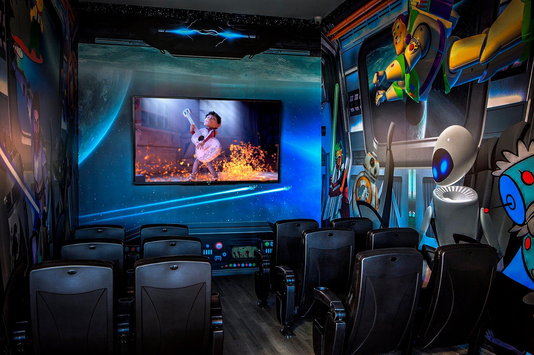 Small theater room with blue-lit walls, rows of seats, and a movie playing on a wall-mounted screen.