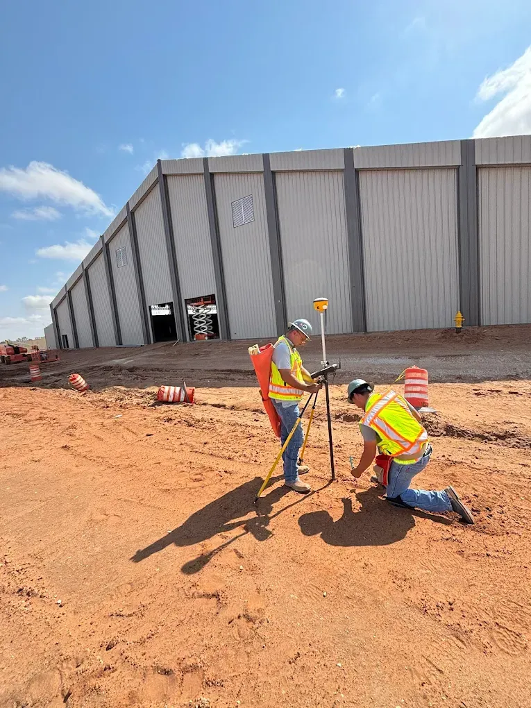 Construction workers measuring dirt beside a large warehouse on a sunny day