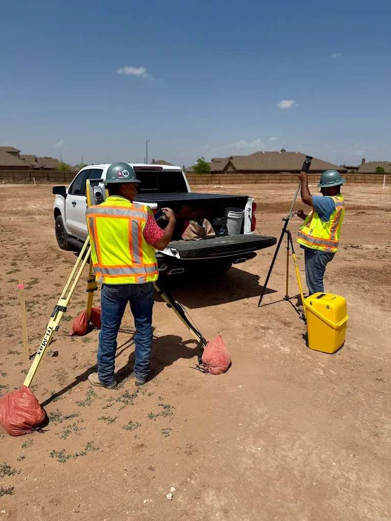 Workers in safety vests set up surveying gear beside a pickup truck on a dusty site.