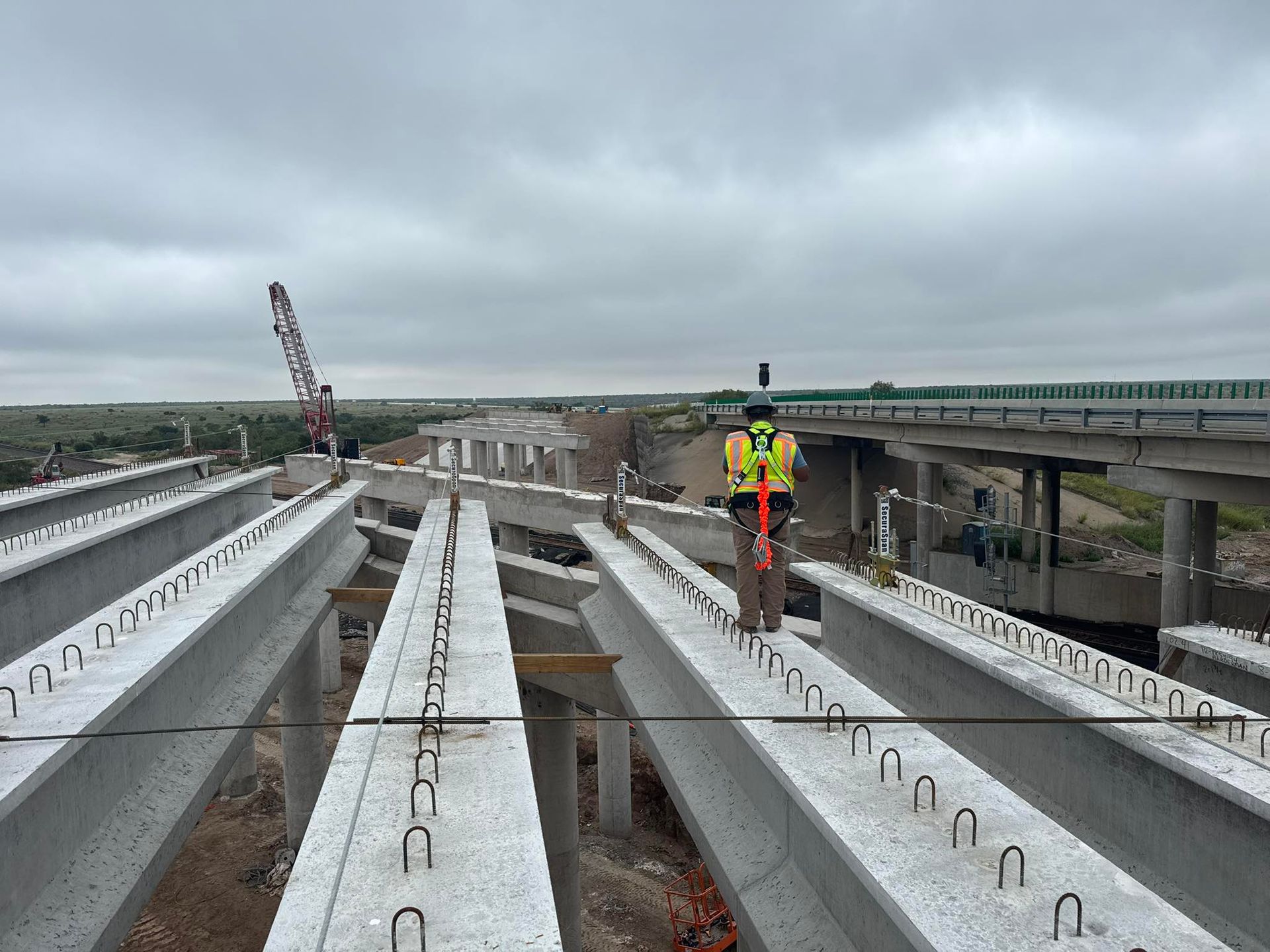 Construction worker on an elevated bridge deck with long gray beams under a cloudy sky.