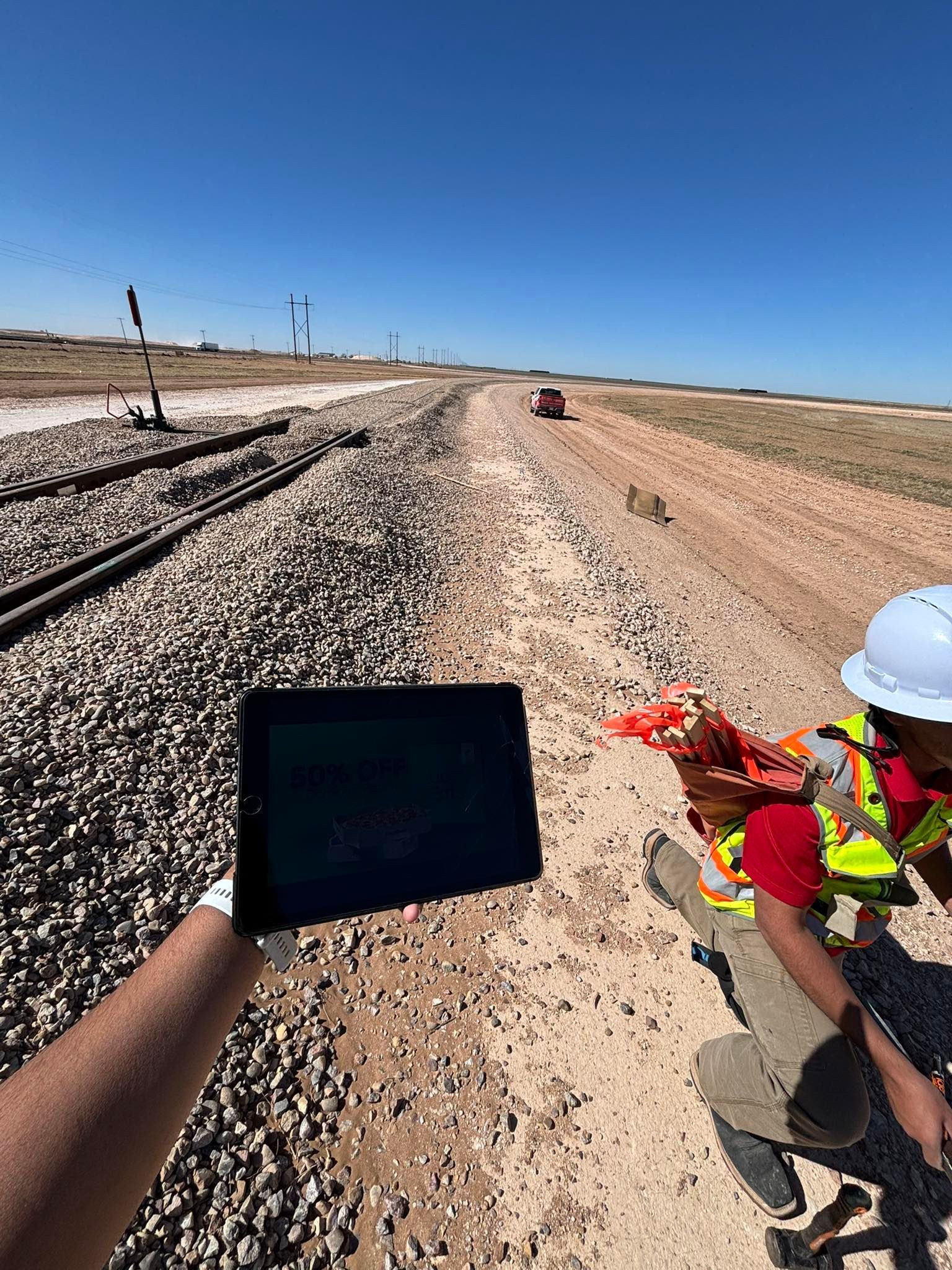 Construction worker beside railroad tracks on a sunny day, holding a tablet and red flowers.
