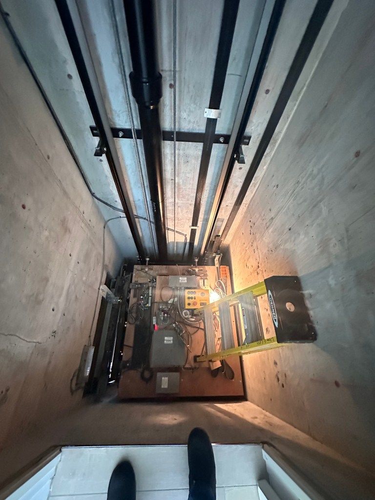 Elevator shaft interior, looking down. Electrical components and cables visible above the platform; concrete walls surround.