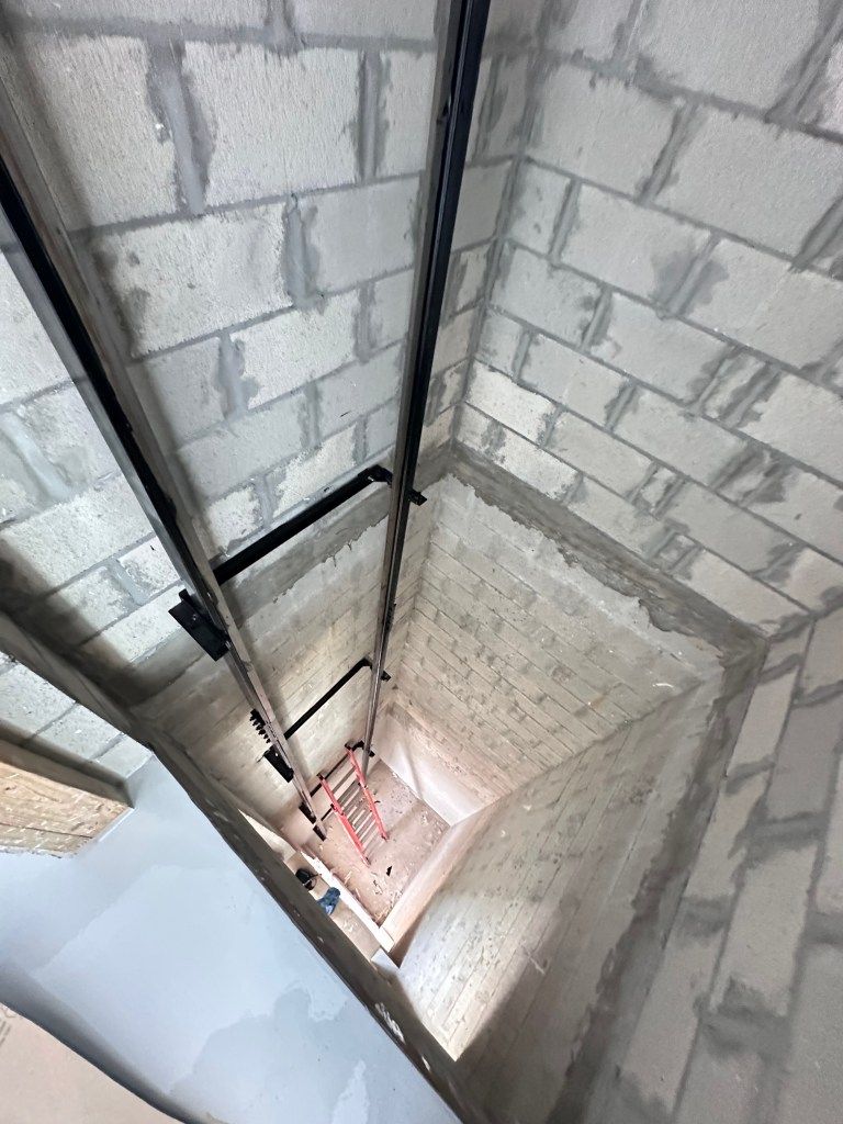 Elevator shaft under construction. Gray cinder block walls. Black rails and ladder visible inside the shaft.