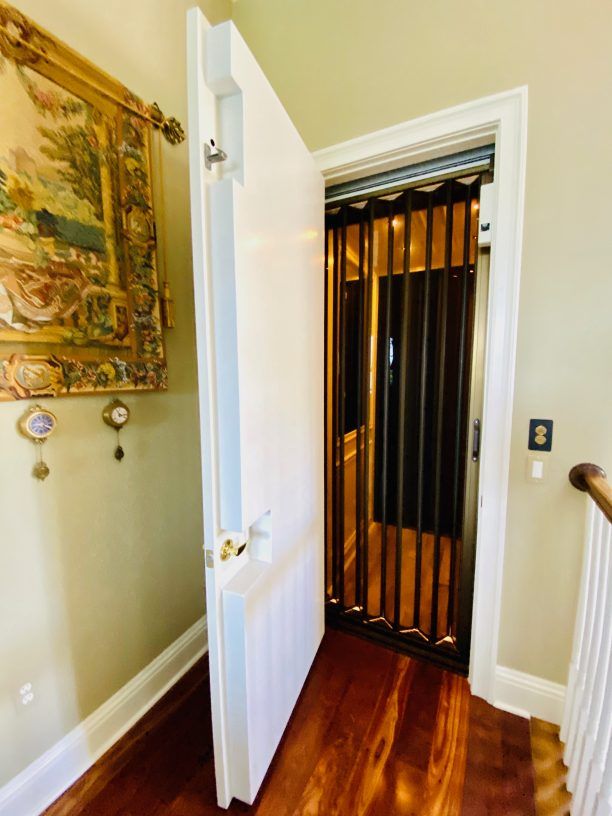 Open white door reveals home elevator. Brown metal gate is visible. Wood floors and off-white walls.