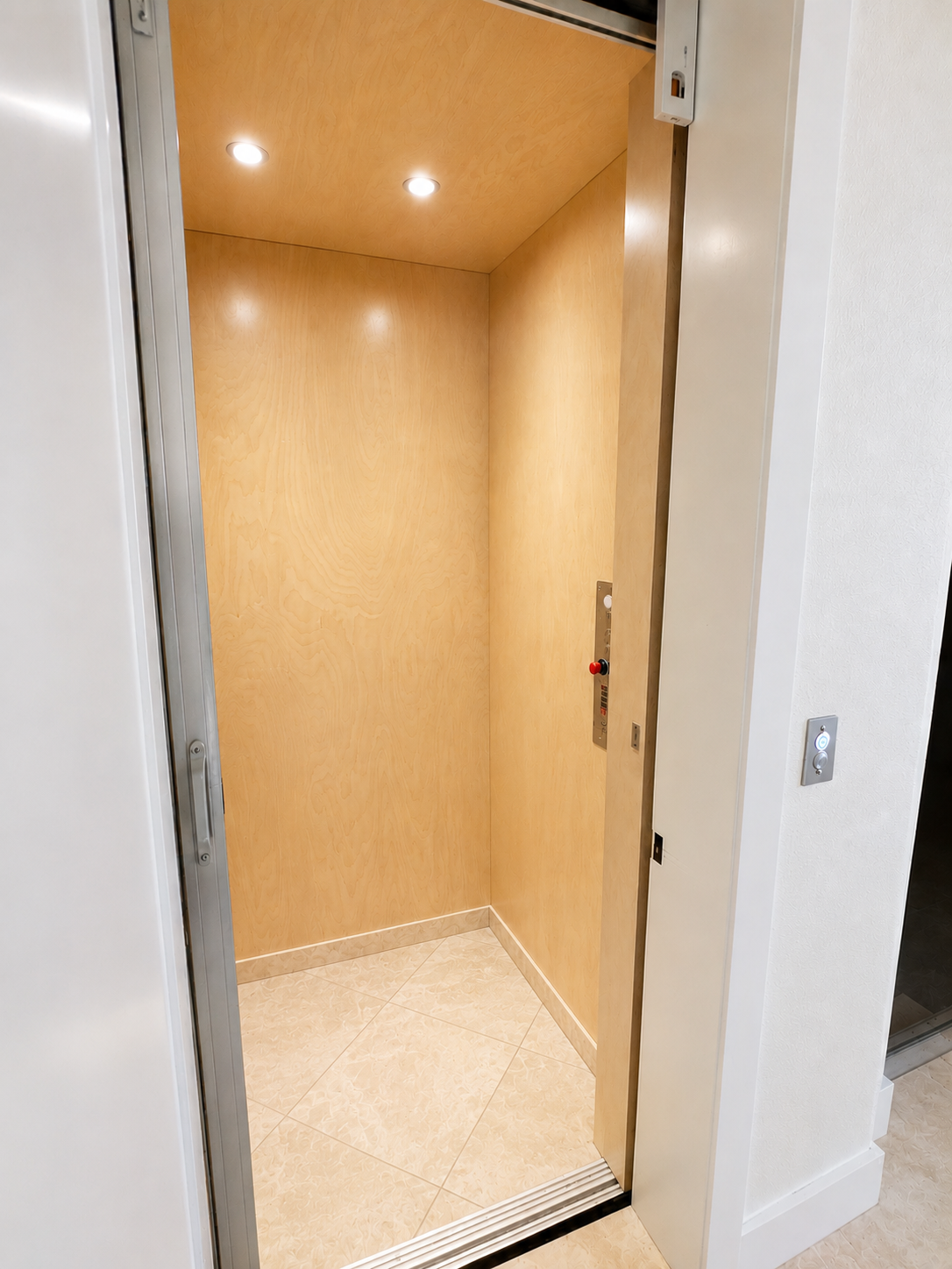 Empty beige elevator with wood-paneled walls and recessed ceiling lights, door open to a white hallway.
