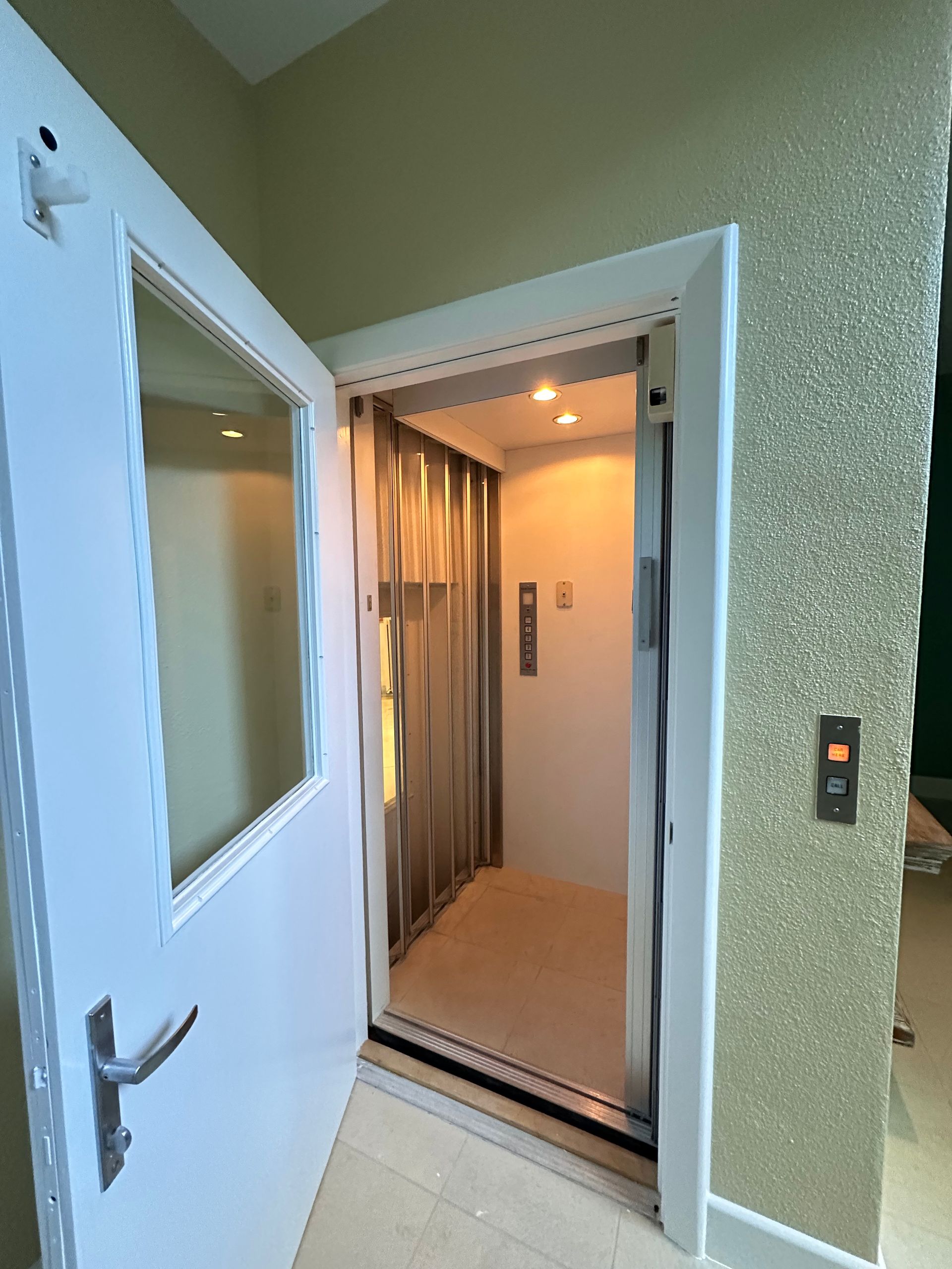 Open elevator doorway, white door with window. Interior: beige walls, control panel, and lights.