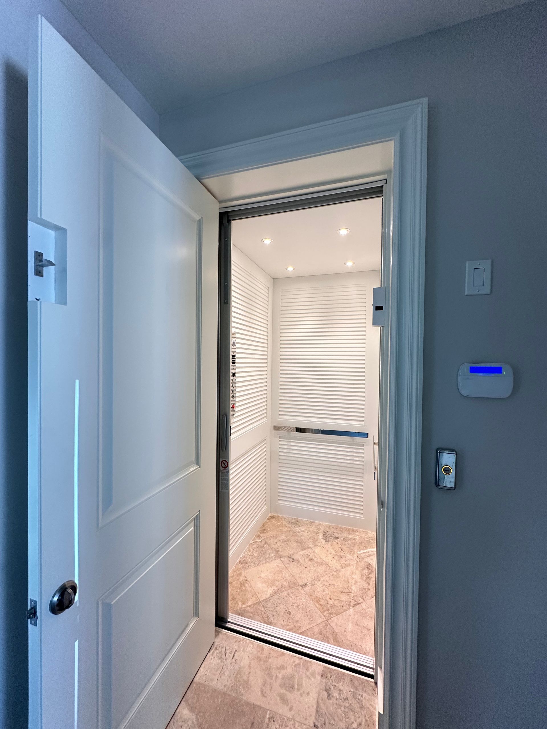 Open doorway revealing an elevator with patterned white interior. The room has light-colored walls and flooring.