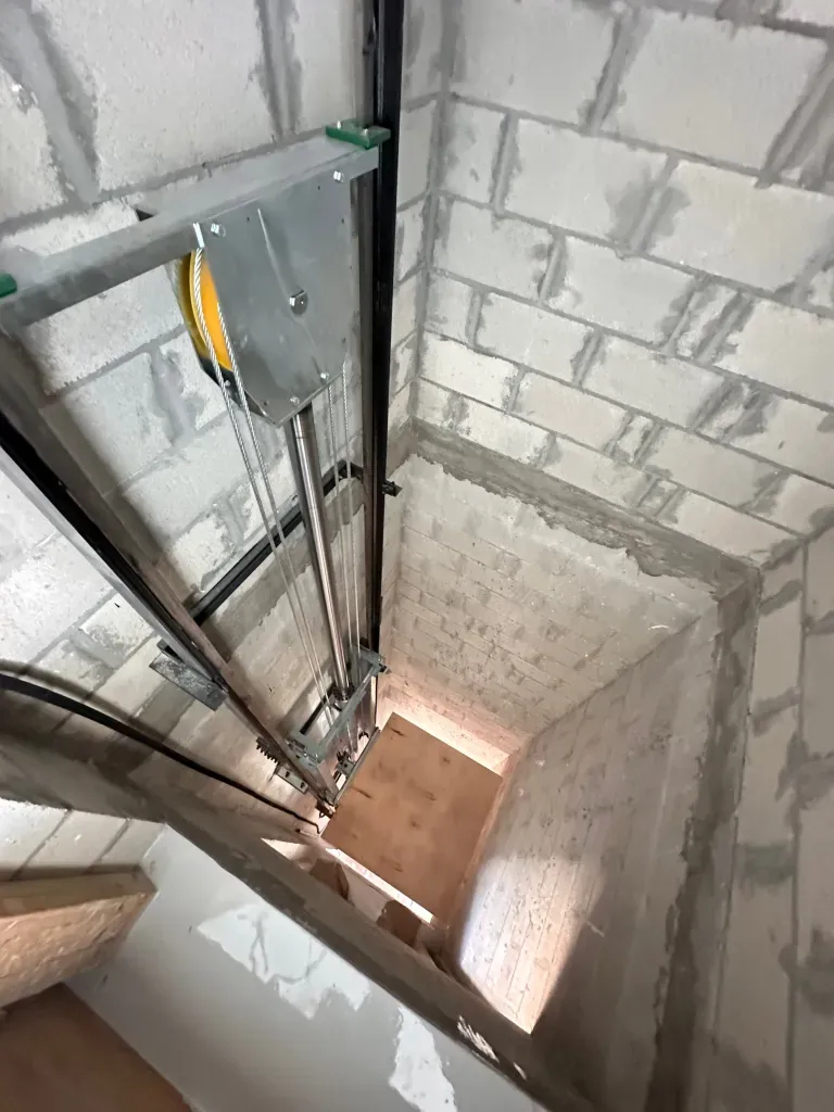 Elevator shaft under construction with exposed machinery and unfinished brick walls, looking down toward stairs.