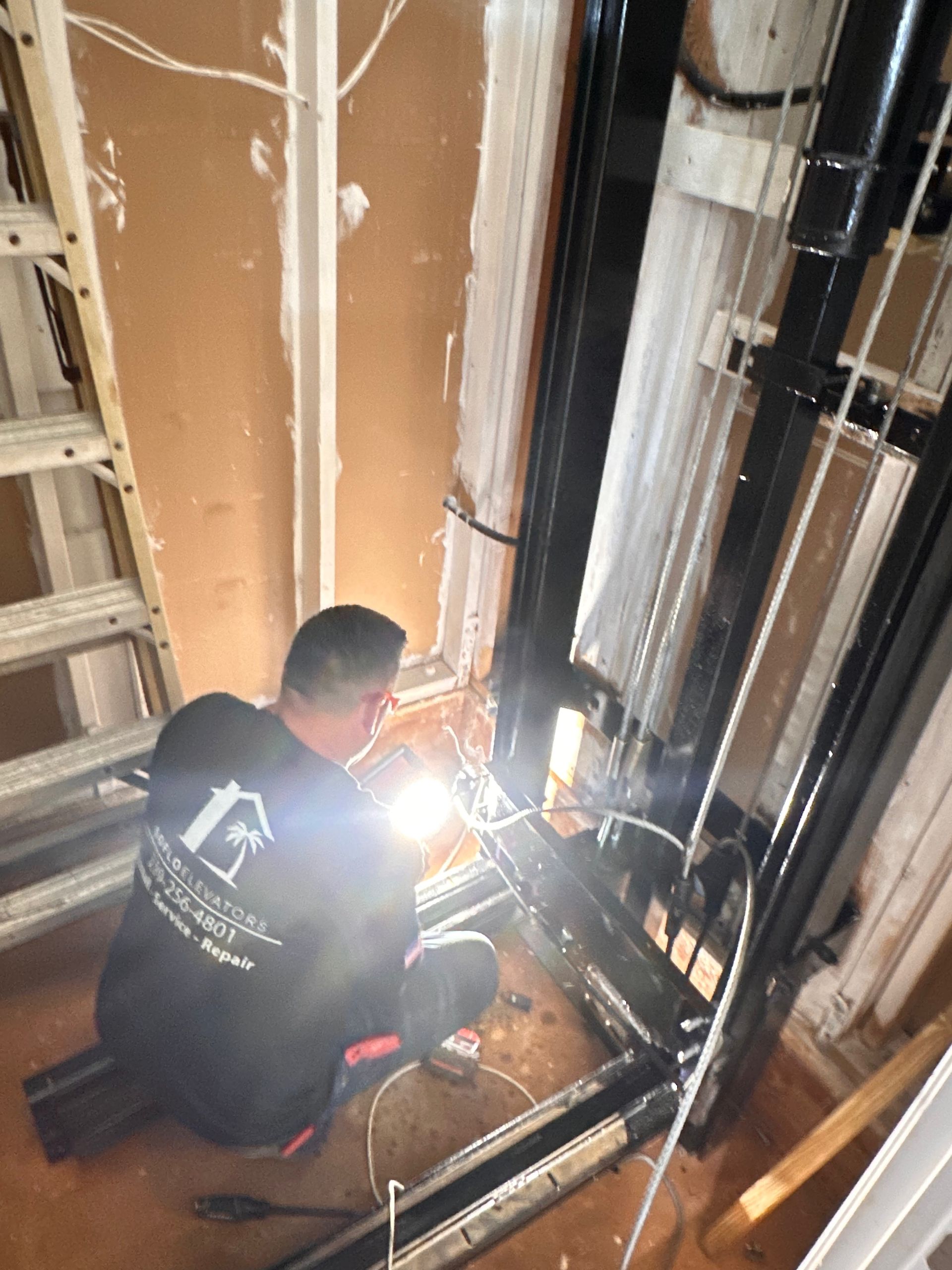 Man working on elevator, crouching with flashlight. Black elevator frame, exposed wiring, inside a building.