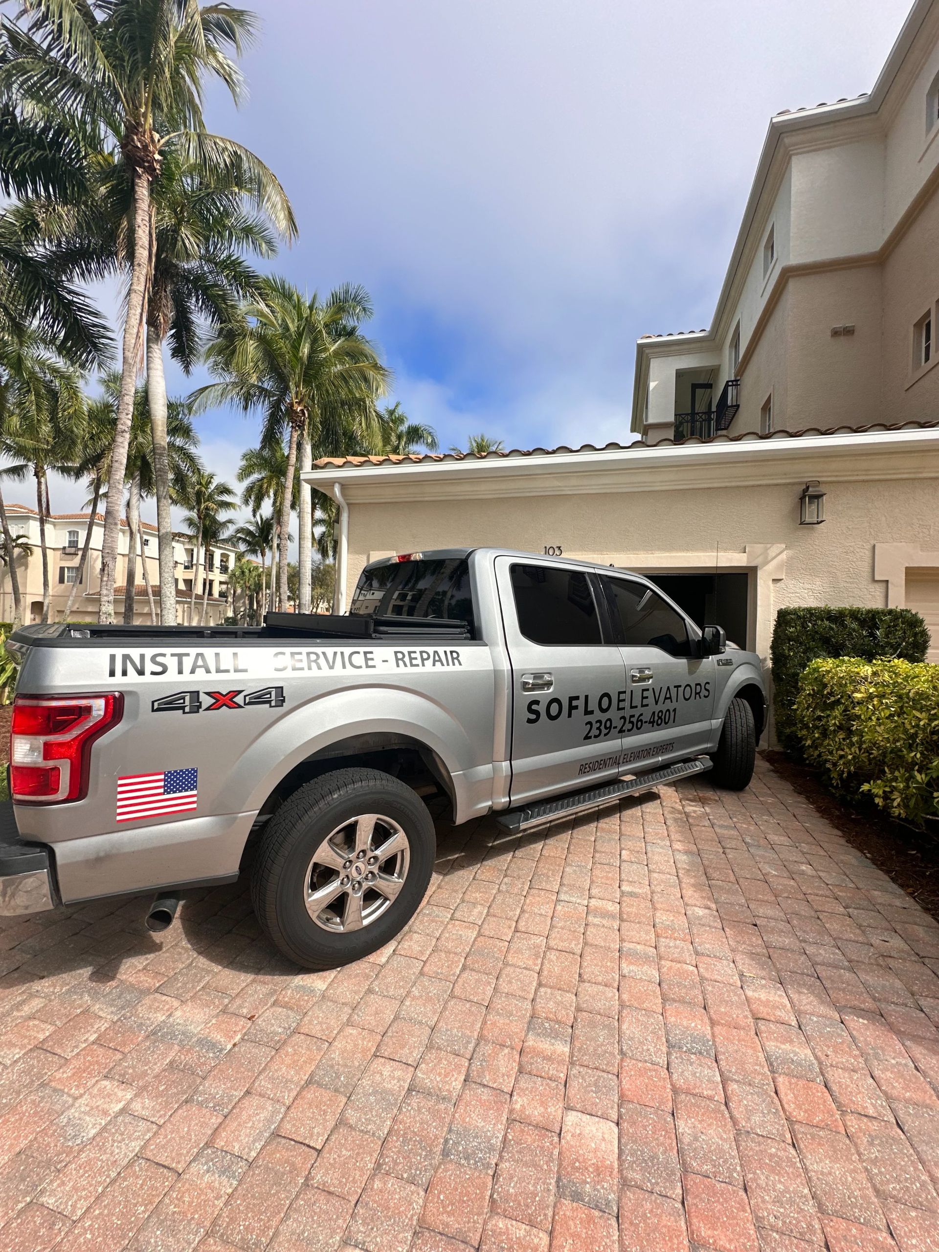 Silver utility truck parked by a beige building in a sunny driveway, with palm trees in the background