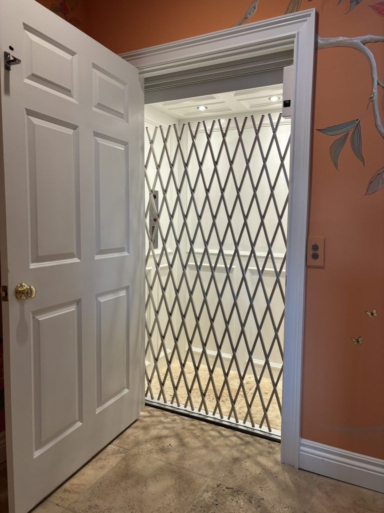 White door opens to an elevator with a diamond-patterned gate inside a room with peach walls and patterned wallpaper.