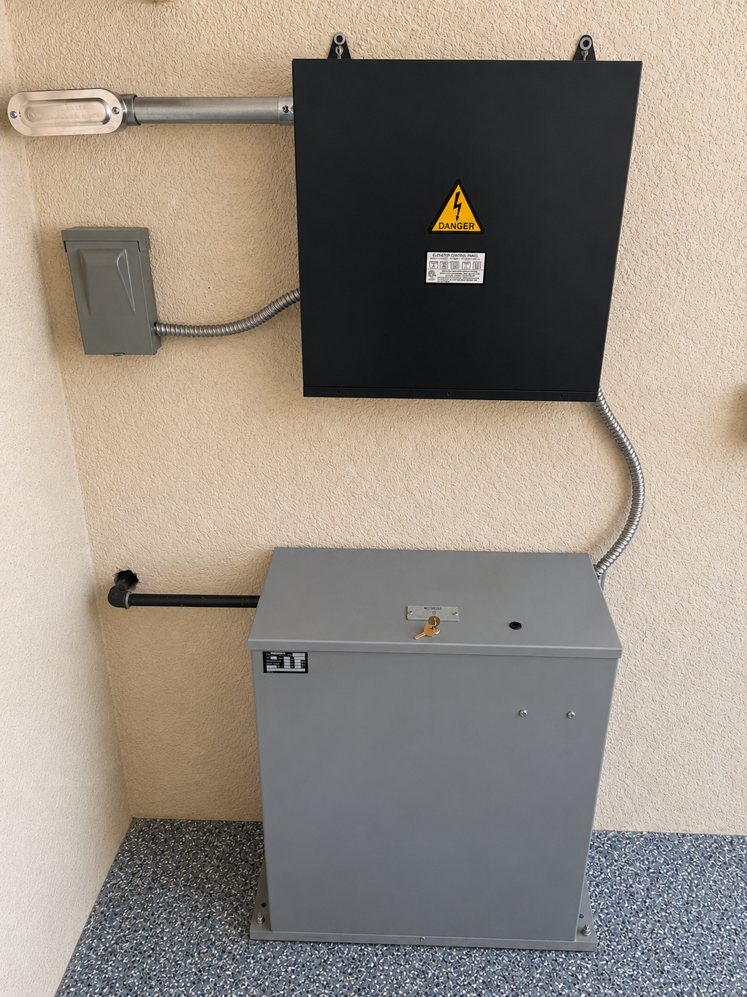 Two utility boxes mounted on a wall, with exposed conduit and a warning symbol on the upper black box.