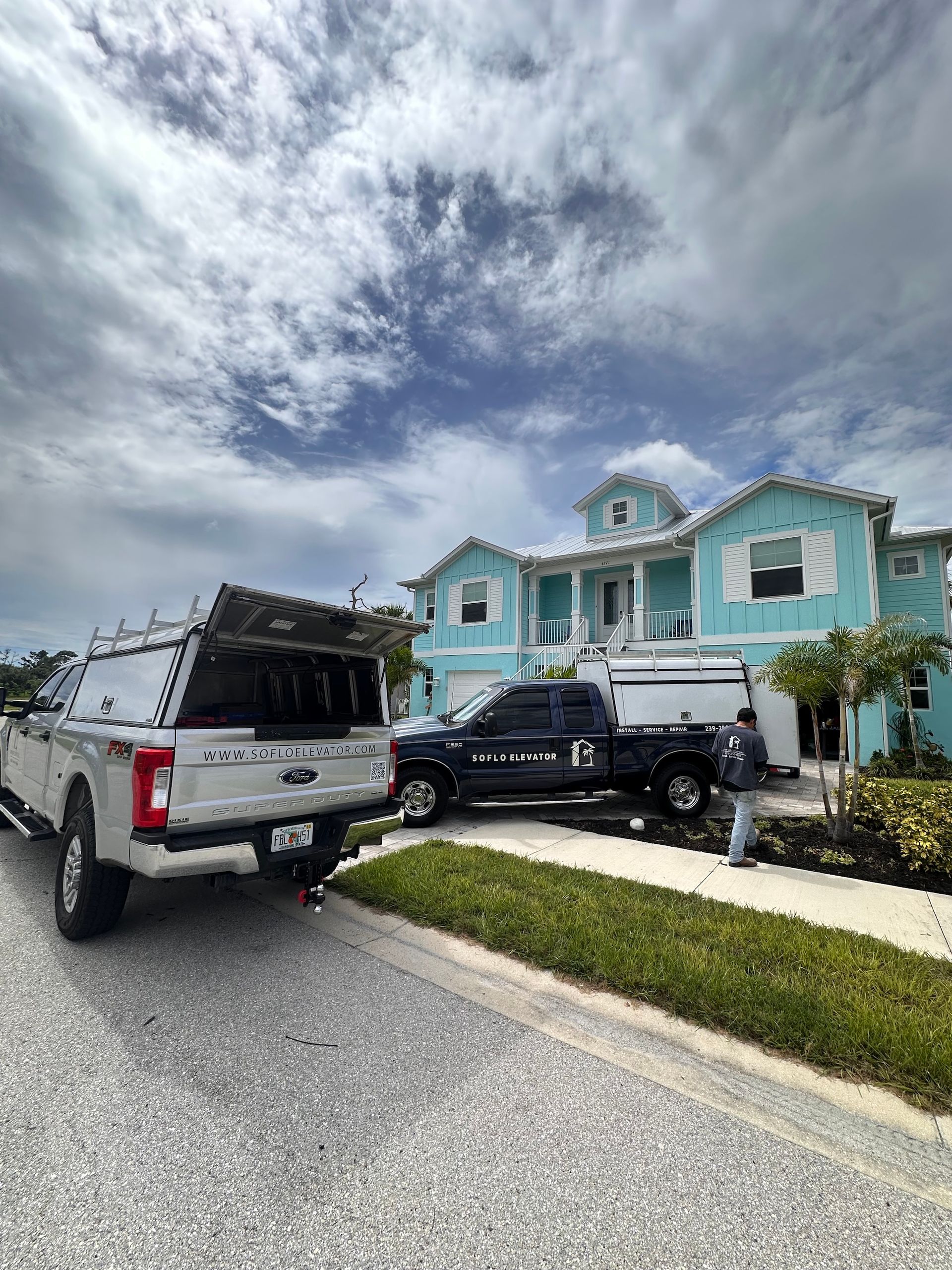 Two trucks parked in front of a blue house with white trim. Man stands near one truck. Overcast sky.