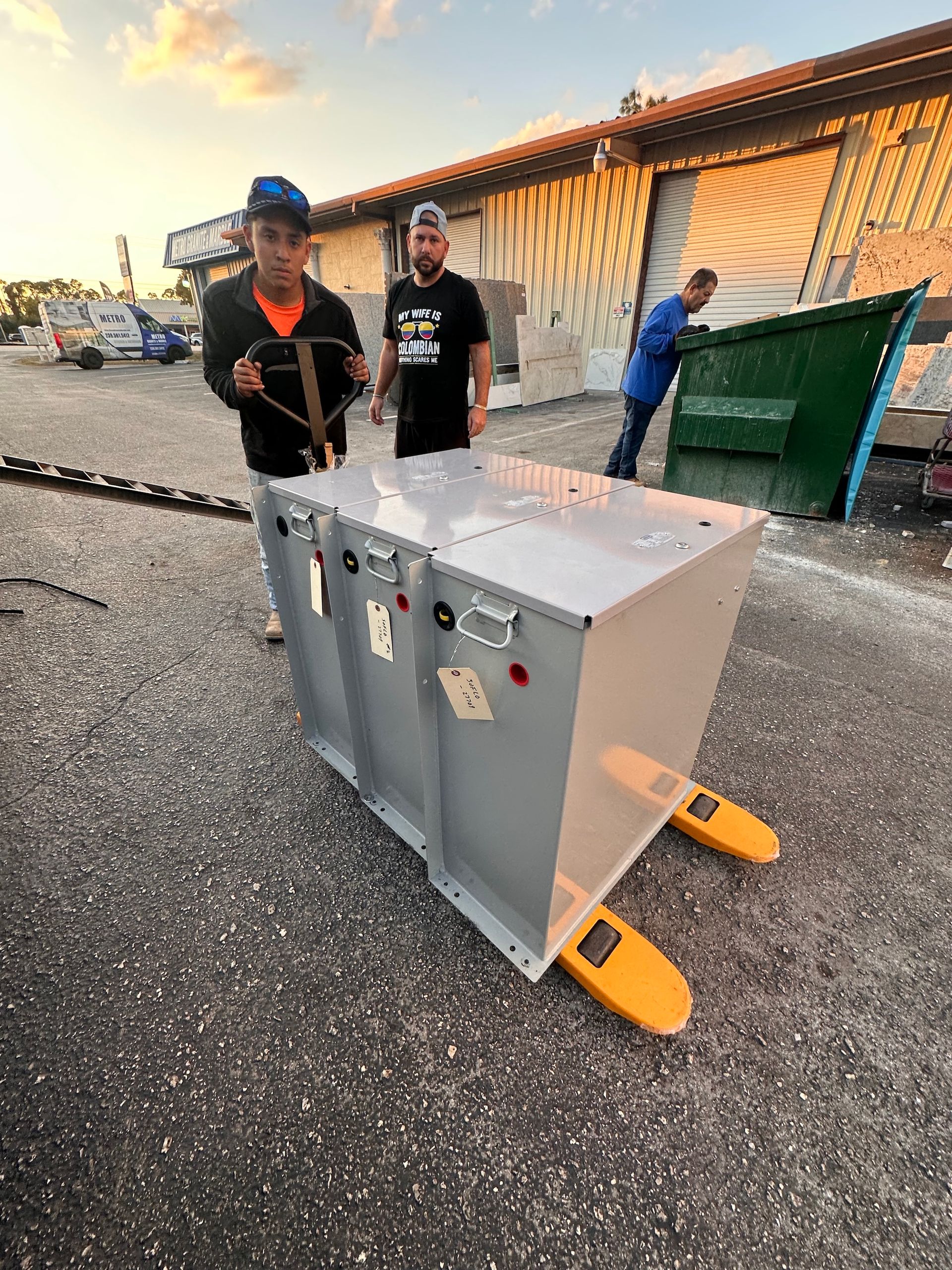 Three people next to a large metal box on a pallet jack outside a building.