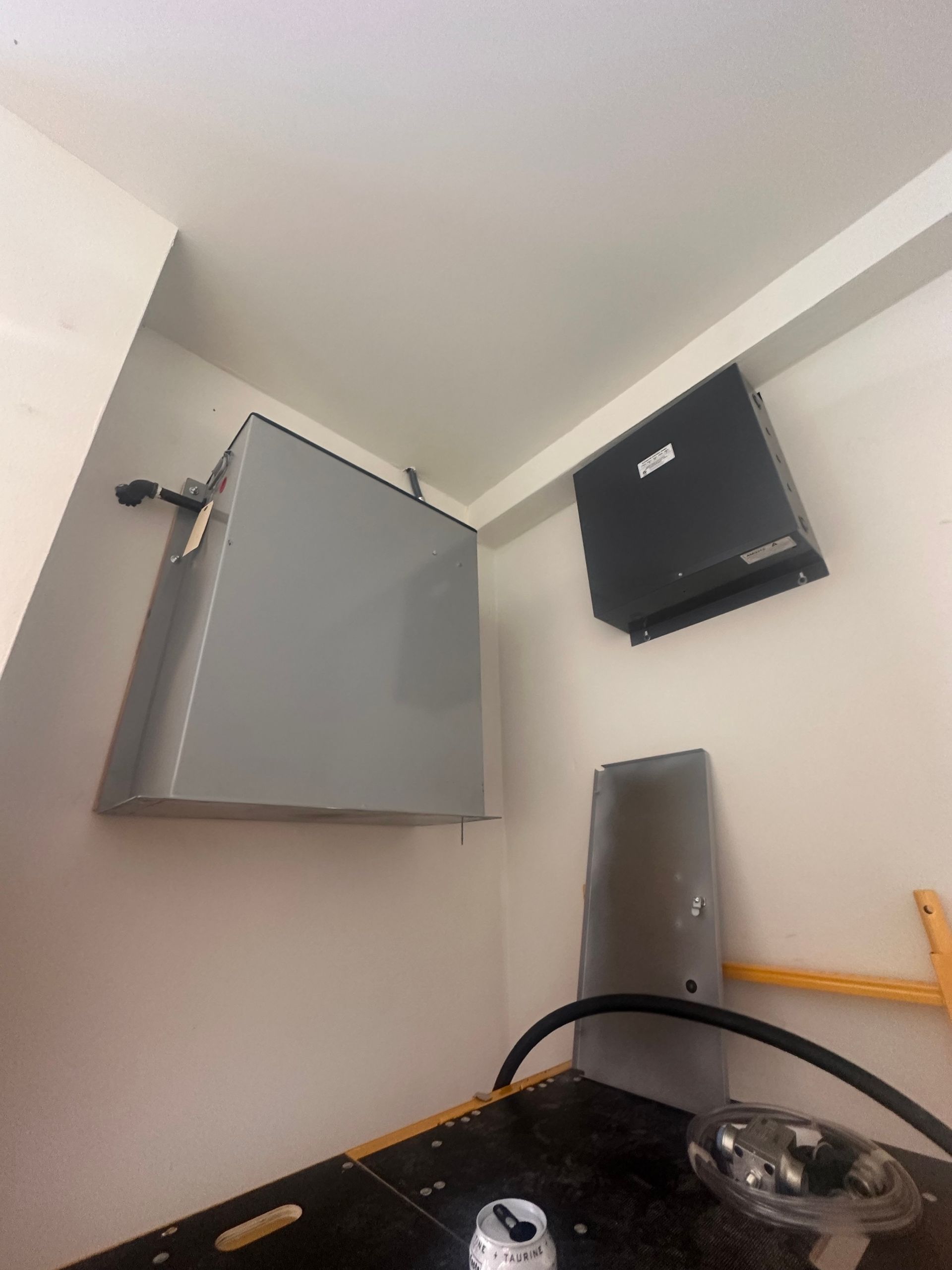 Gray metal electrical box and black panel mounted on a white wall in a corner.