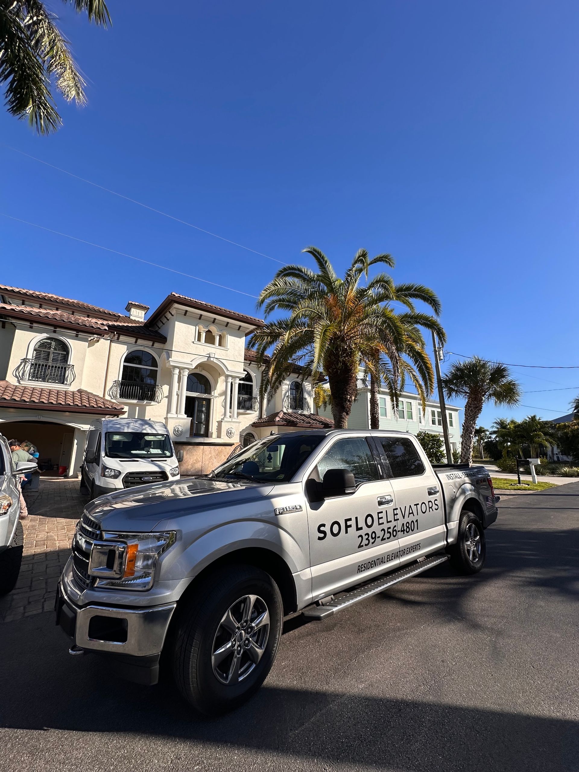 Silver pickup truck parked in front of a large house with palm trees, under a blue sky.