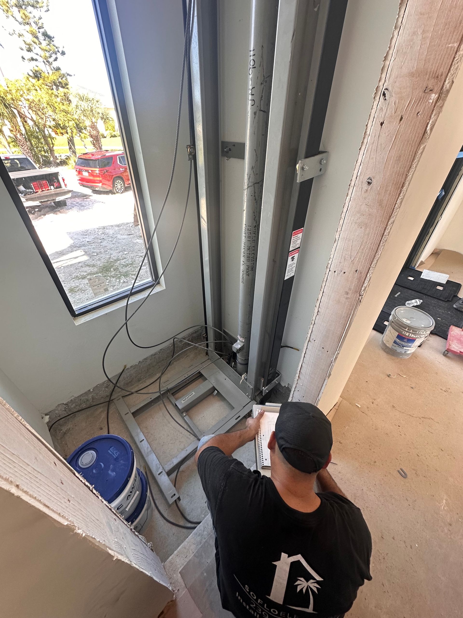 Man inspecting a home lift installation near a window. Gray metal lift structure, concrete floor, white walls.