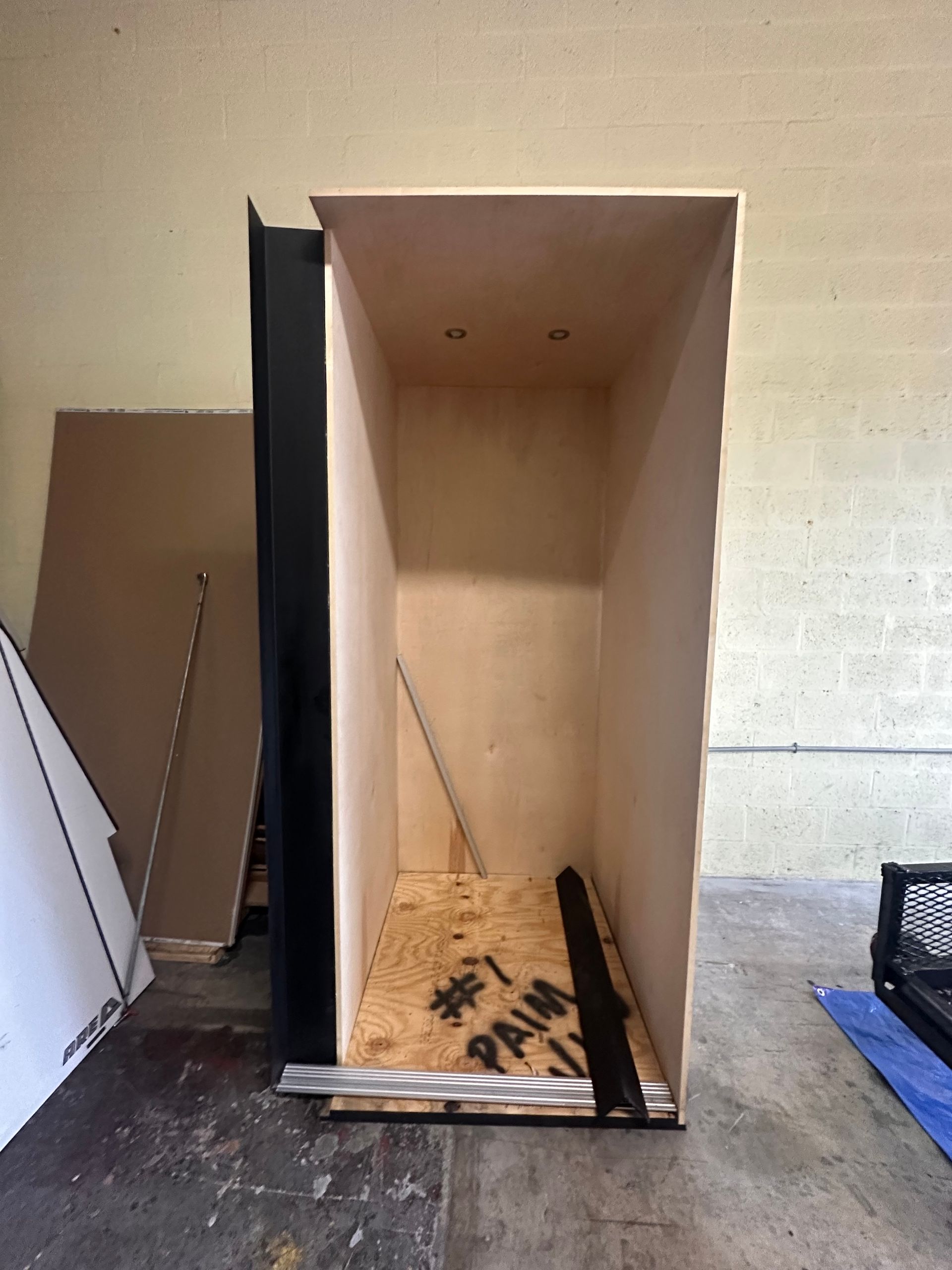 Interior view of a plywood box with a black frame and unfinished wood base in a workshop.