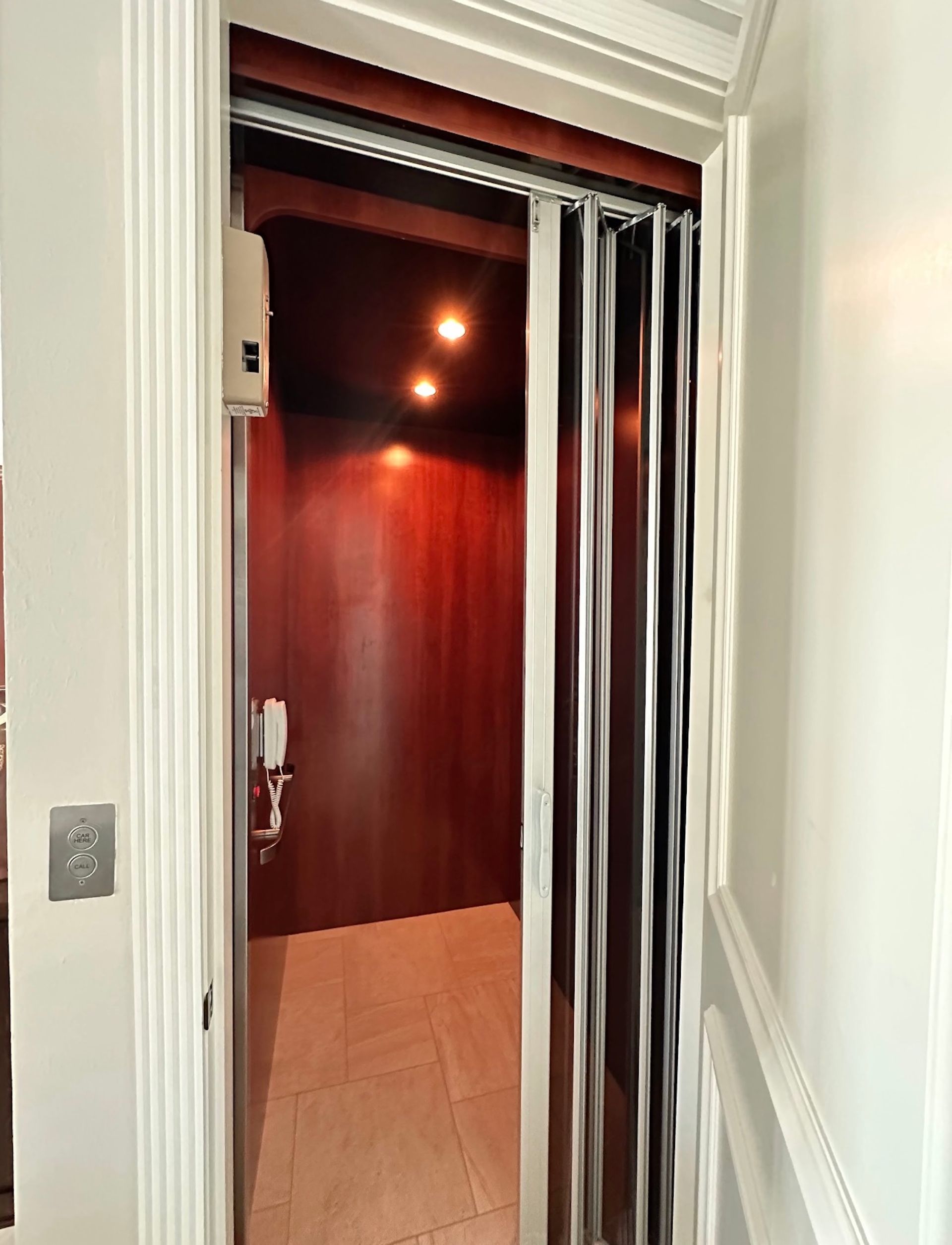 Elevator with accordion door partially open. Dark wood interior, overhead lights, wooden floor, and wall phone.