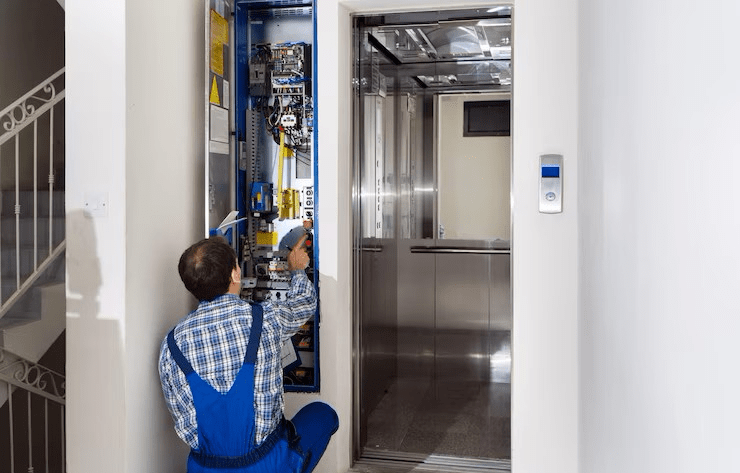 Elevator technician in blue overalls working on control panel. Elevator car open, hallway setting.
