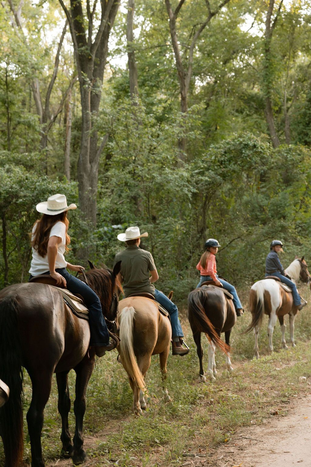 People horseback riding along a forest trail in hats and casual clothes
