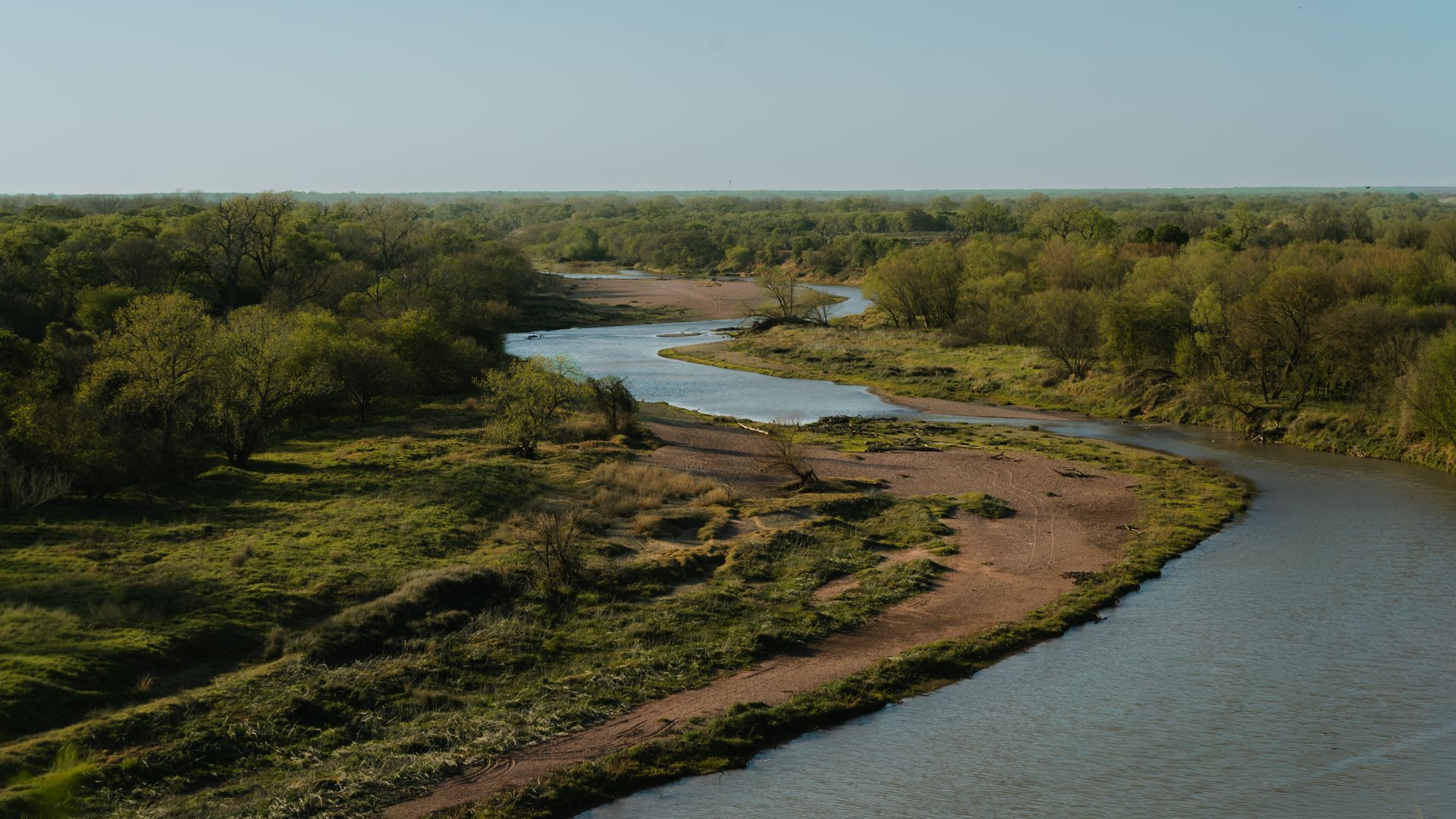 Winding river through green wetlands under a clear sky, viewed from above.