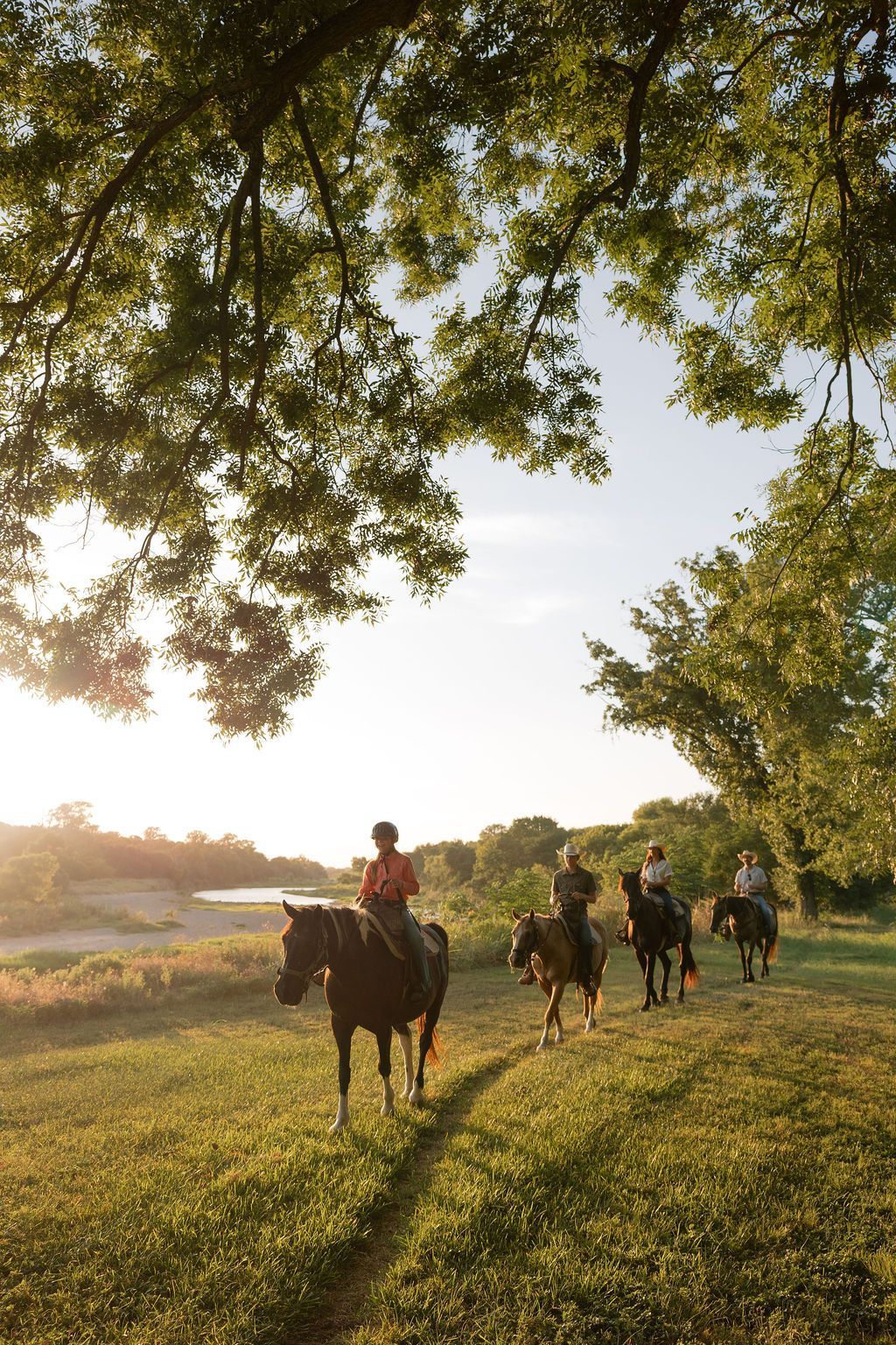People horseback riding in a field, with trees in the background under an overcast sky.