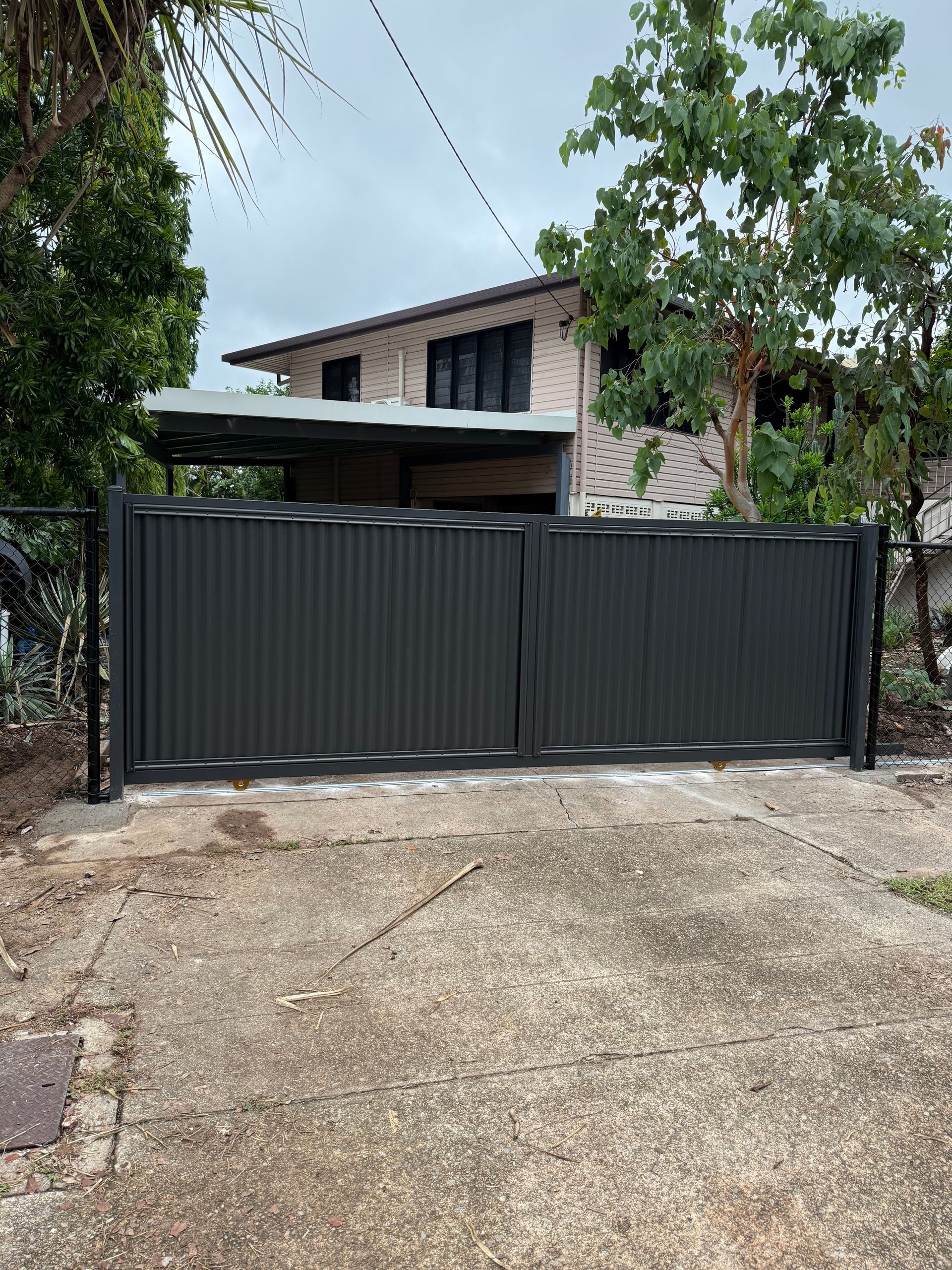 Black corrugated metal gate in front of a two-story house with trees and overcast sky. — True North Fencing in Noonamah, NT