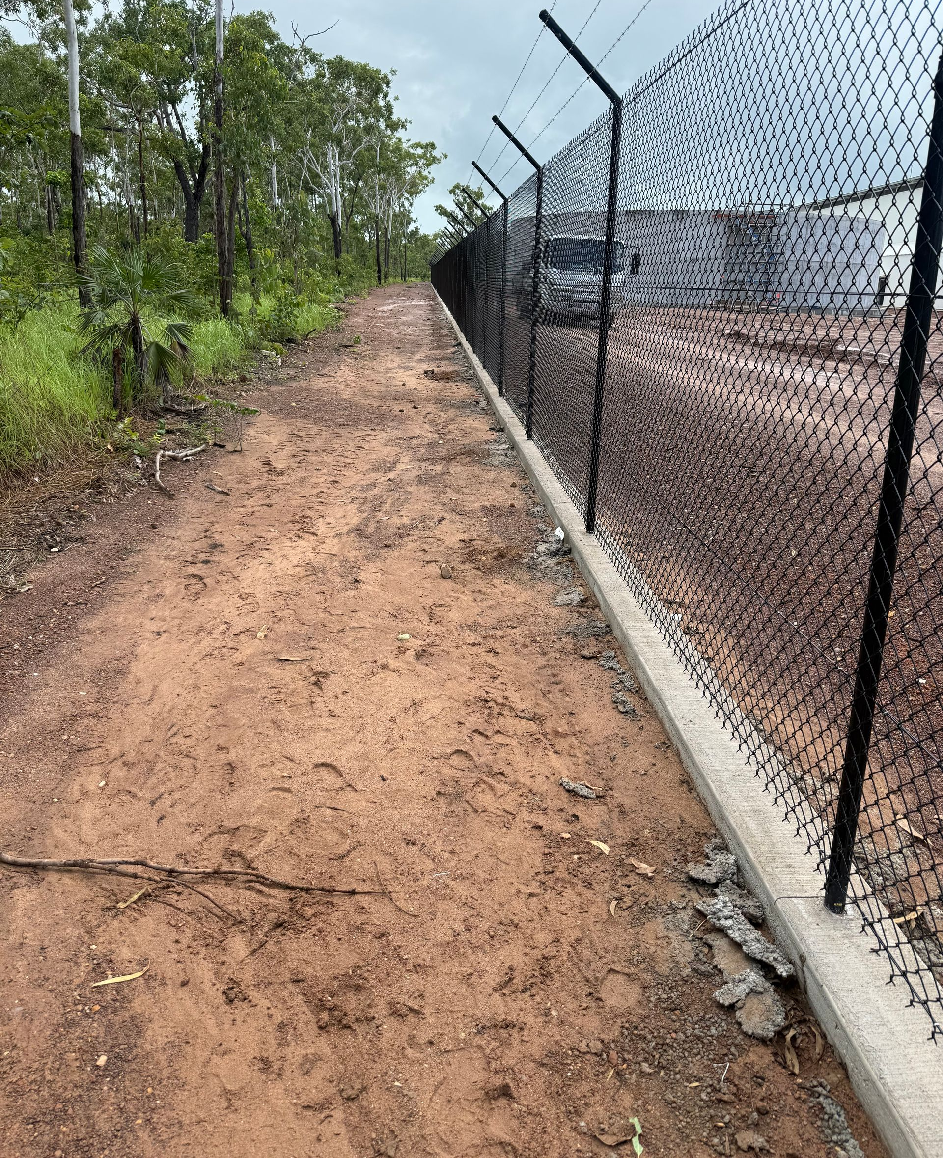 Dirt path alongside a black chain-link fence topped with barbed wire; trees on the left. — True North Fencing In Noonamah, NT