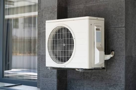 Wall-mounted outdoor heat pump on a dark tiled building wall beside a window