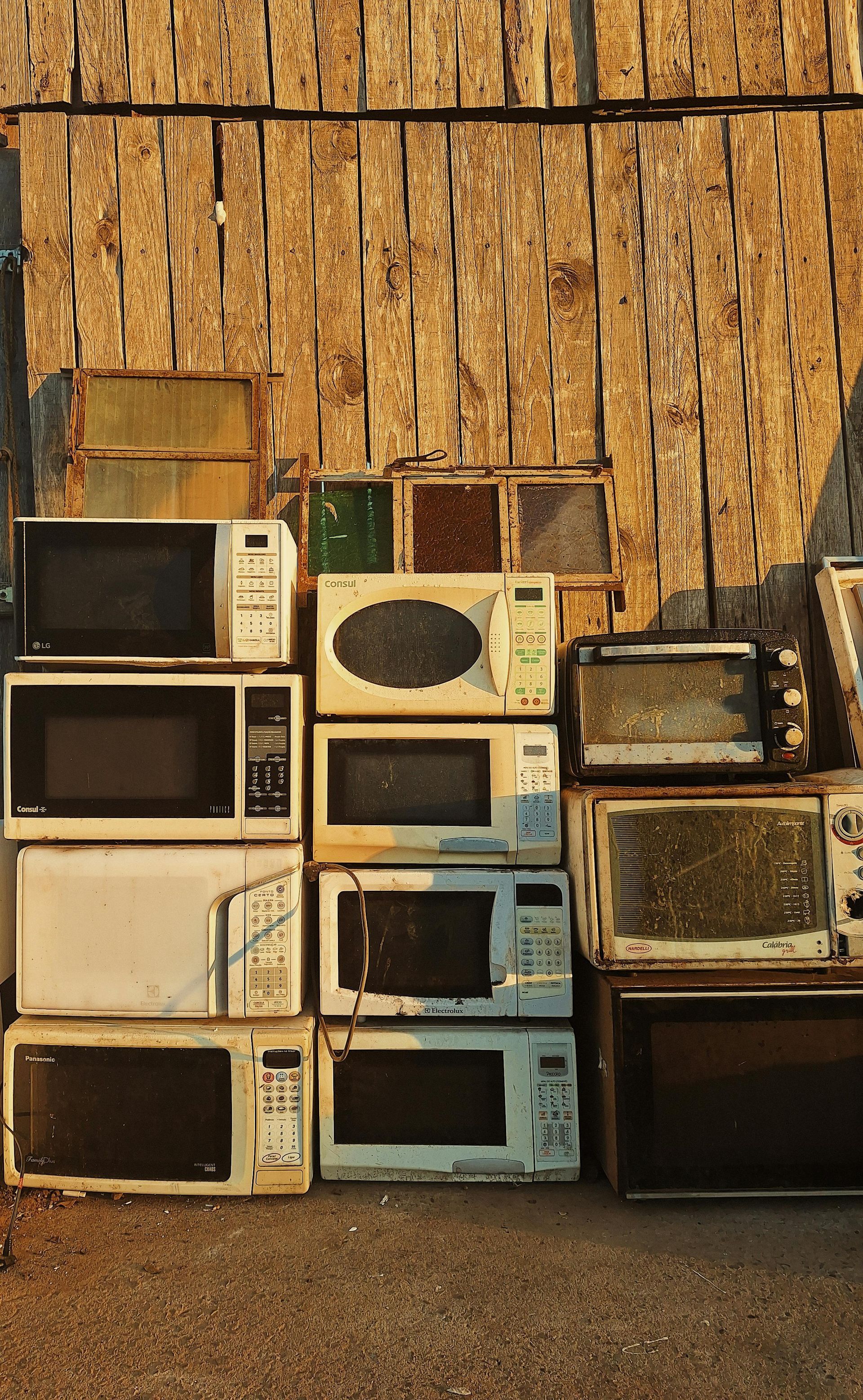 Stack of old microwaves and small appliances against a straw-textured wall.