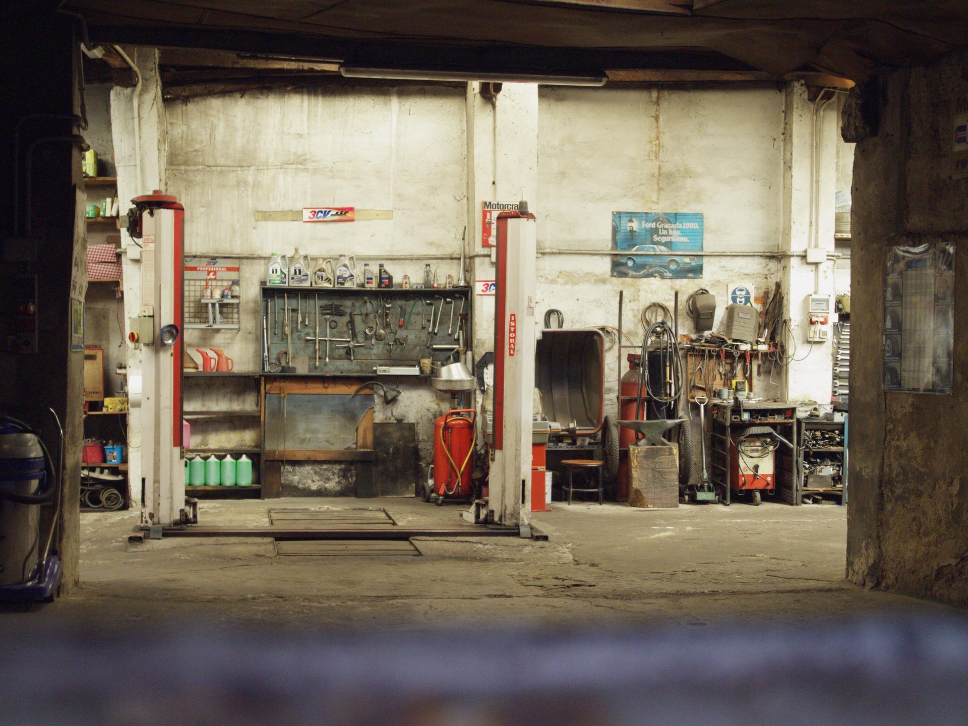 Dim industrial workshop with tools, shelves, and equipment against a worn white wall