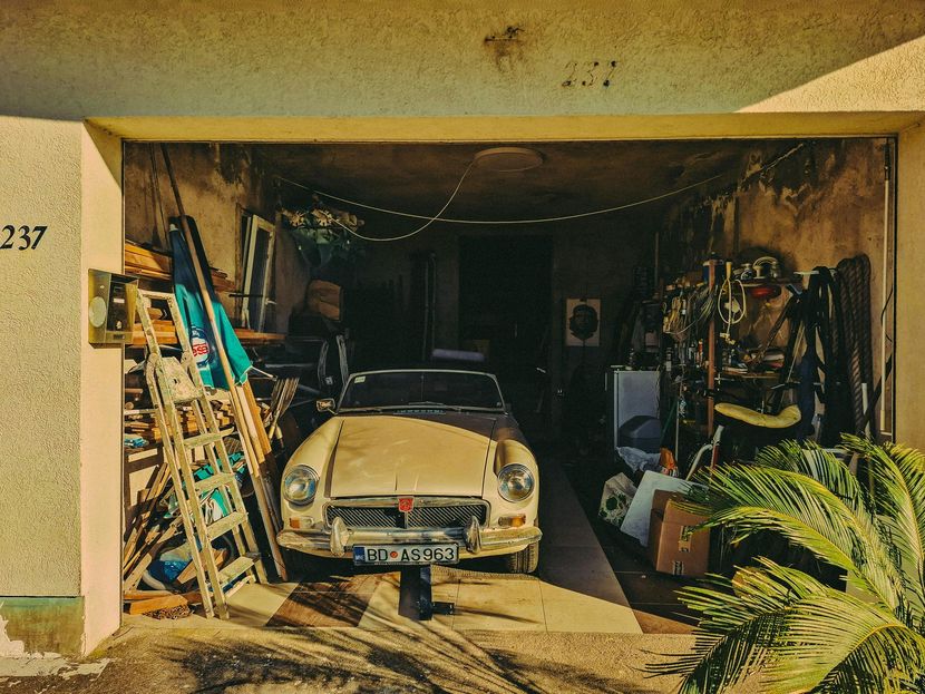 Old beige car parked in a cluttered garage with tools, ladders, and plants around it Garage cleanout service Sacramento CA.