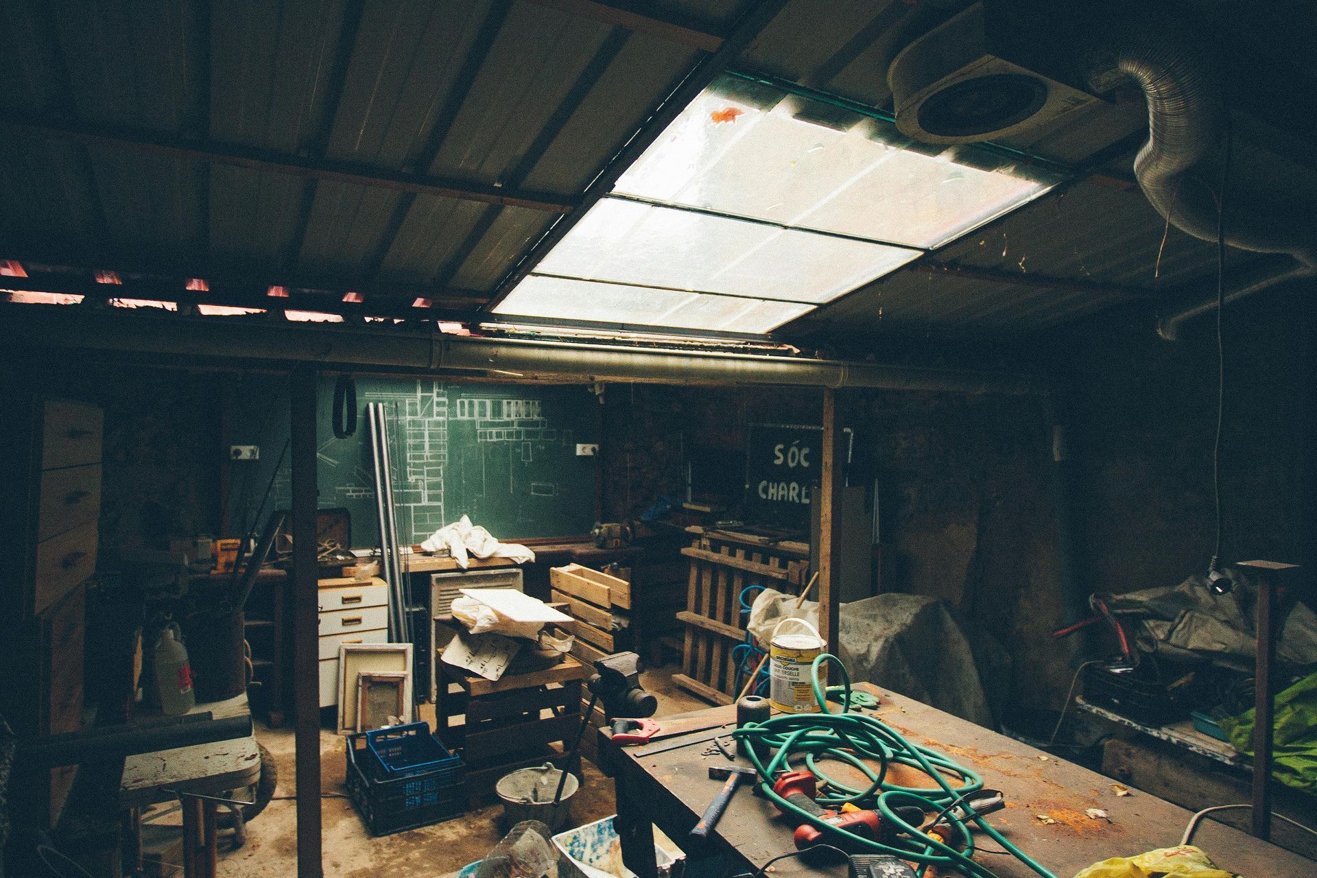Dim cluttered workshop with a corrugated roof, shelves, tools, and a green hose on a workbench.