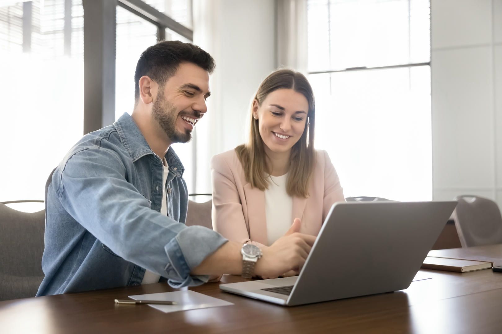 Man and woman smiling at a laptop in a brightly lit office, collaborating on a project.