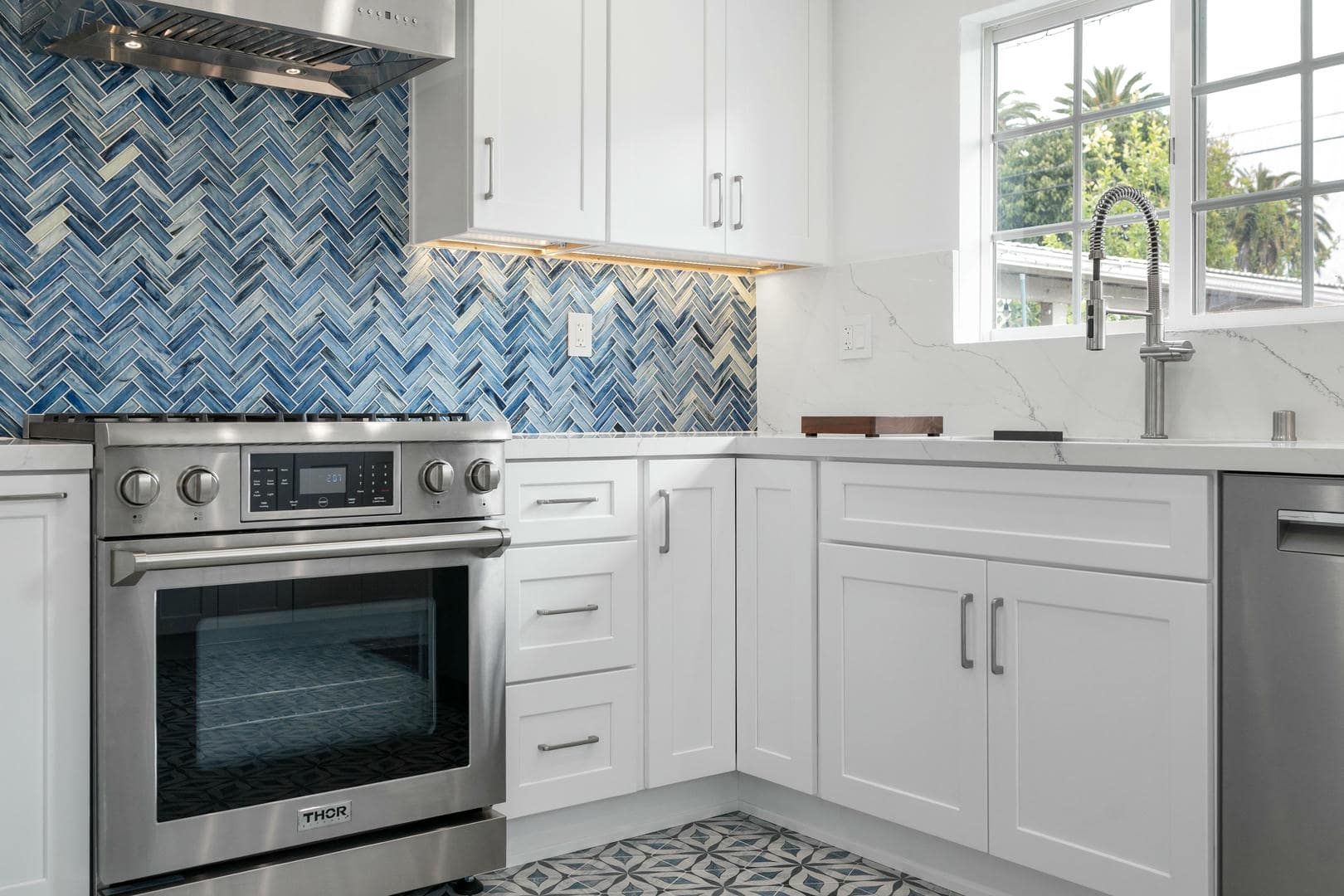 White kitchen with blue herringbone backsplash, stainless steel appliances, and a window.