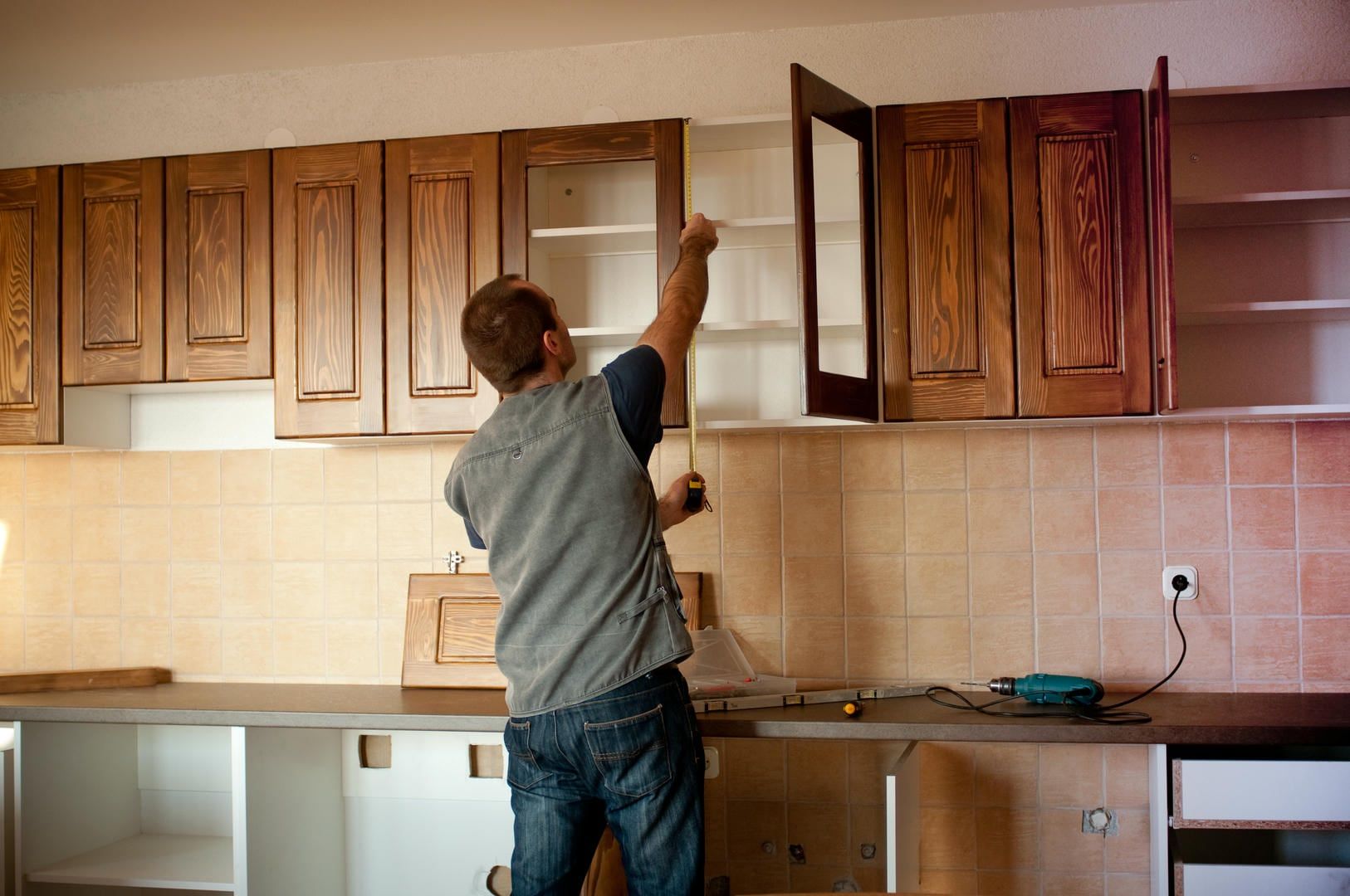 Man installing cabinet in kitchen; brown cabinets, beige backsplash.