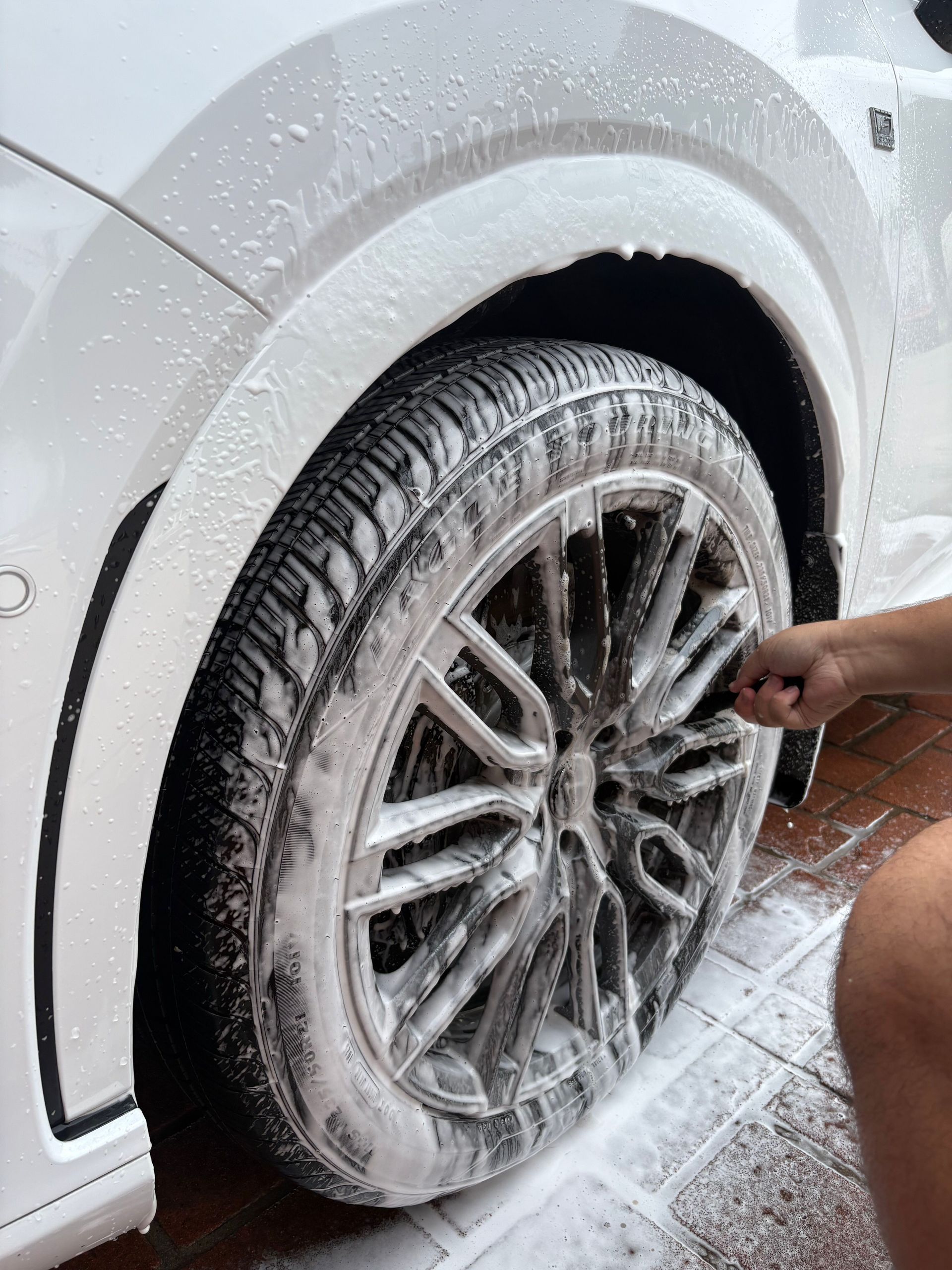Person hand-washing a white car’s wheel with soapy foam at a car wash.