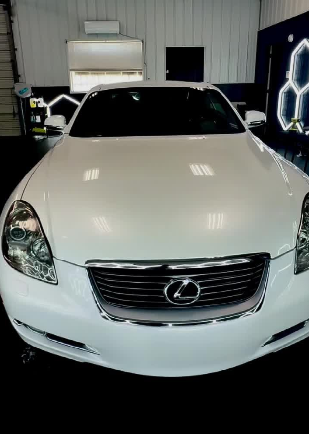 White Lexus sedan parked indoors under bright lights, front view with dark tinted windshield.