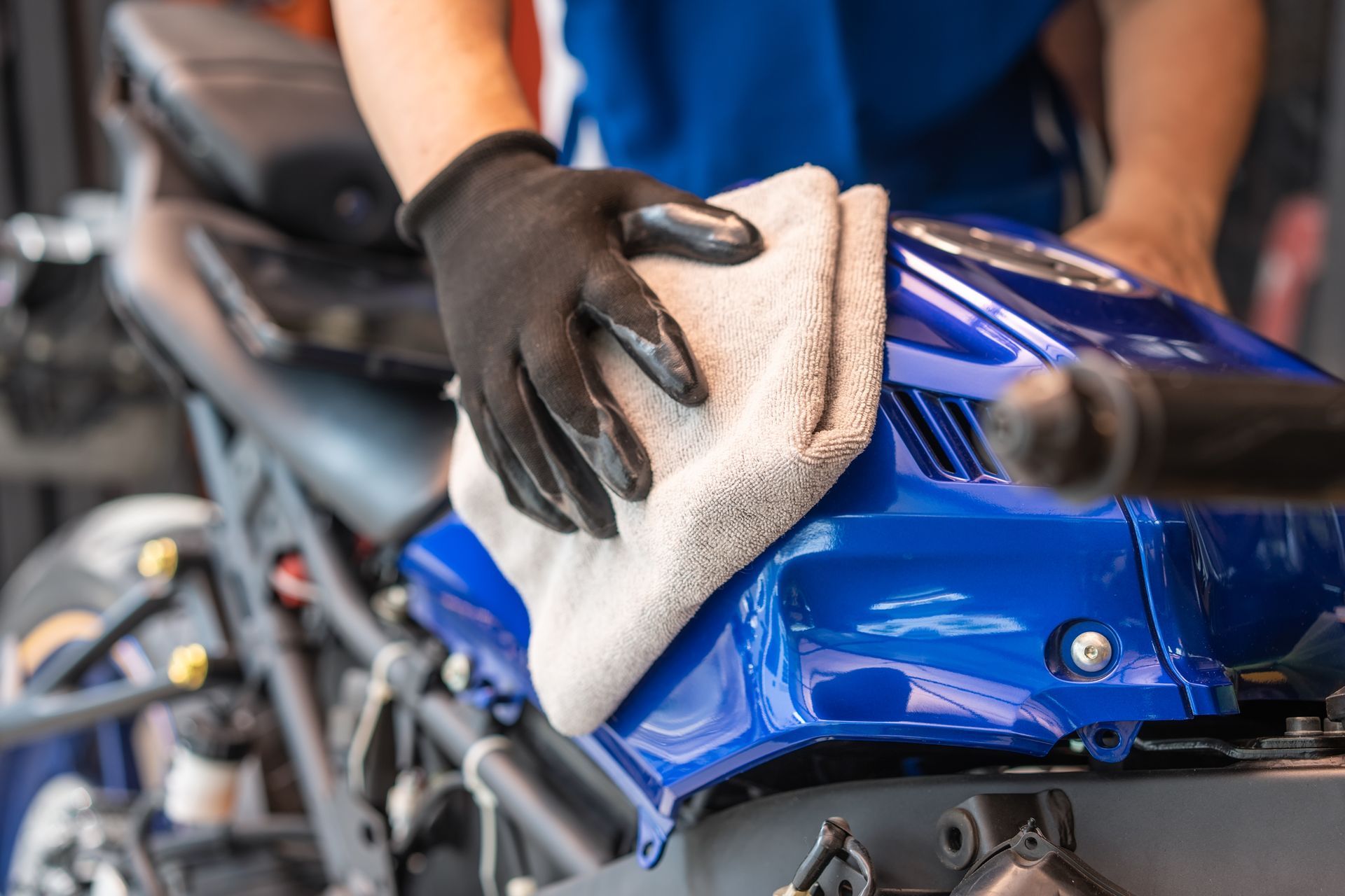 Gloved hands wiping a blue motorcycle with a microfiber cloth in a workshop