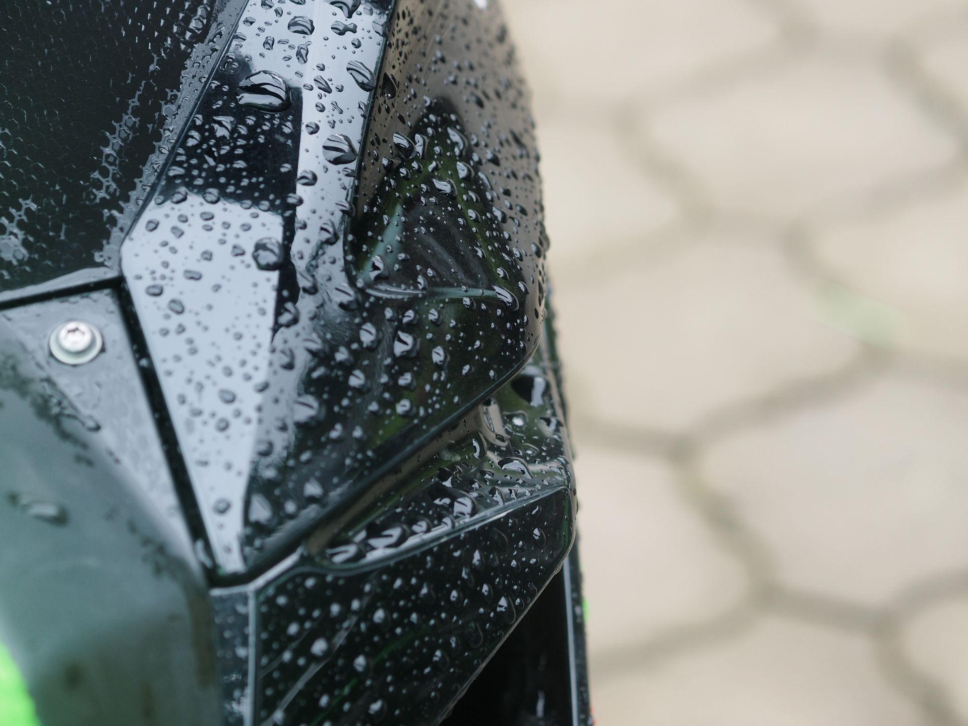 Rain-speckled black car side mirror with green body panel and tiled pavement in the background