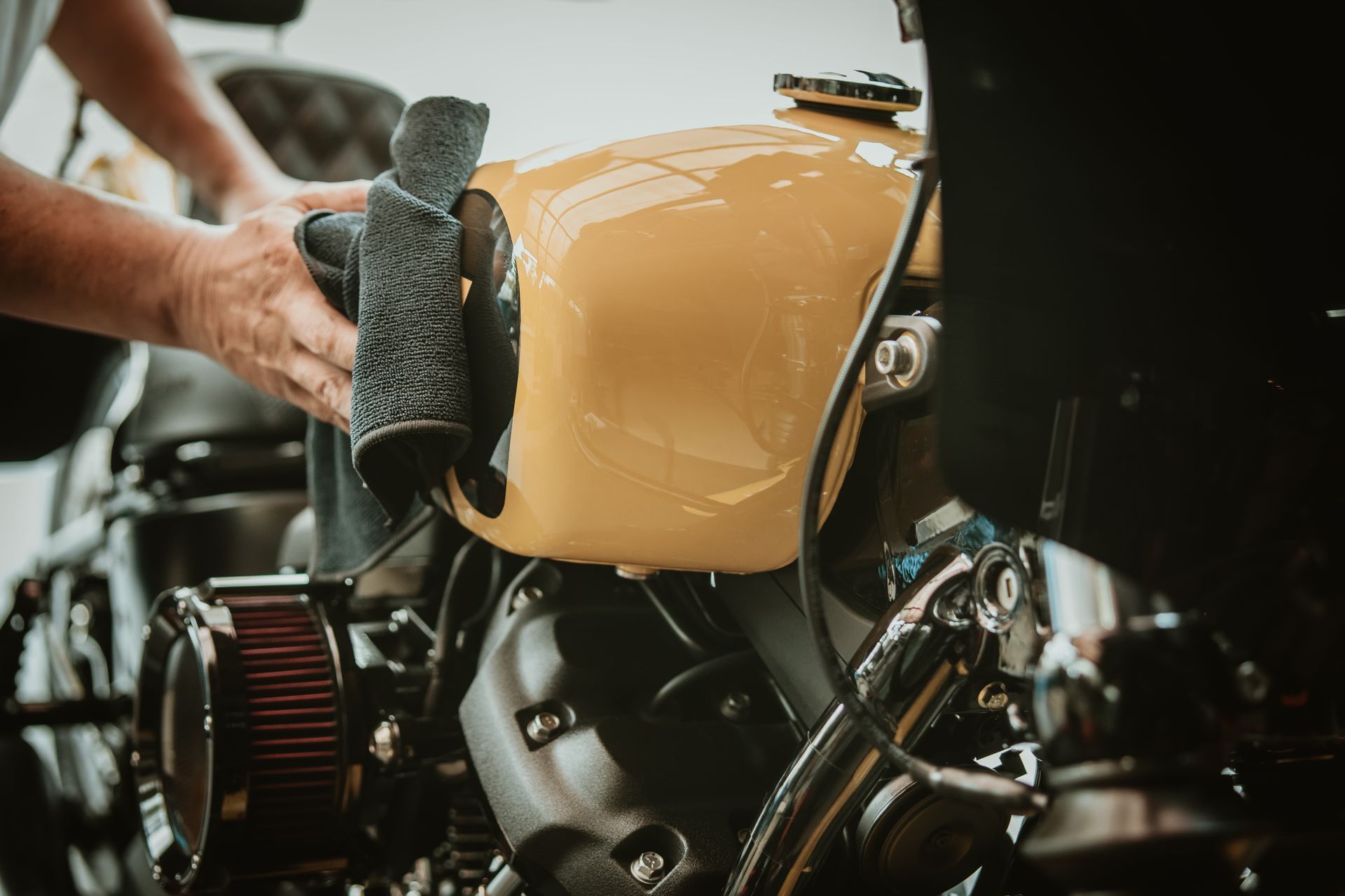 Close-up of a tan motorcycle fuel tank and engine, with a mechanic’s hand working nearby.