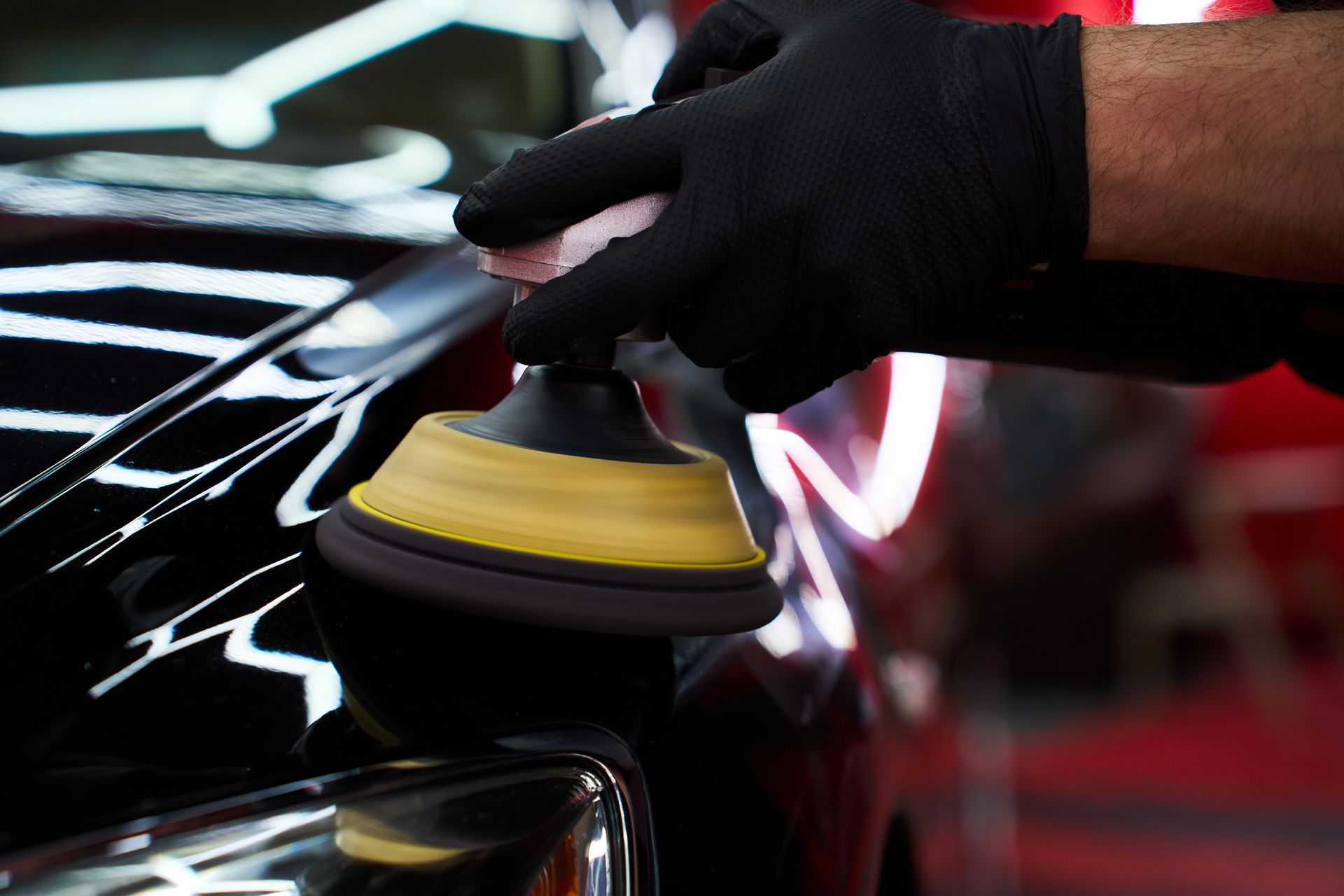 Gloved hand polishing a black car with a yellow buffer pad in a garage