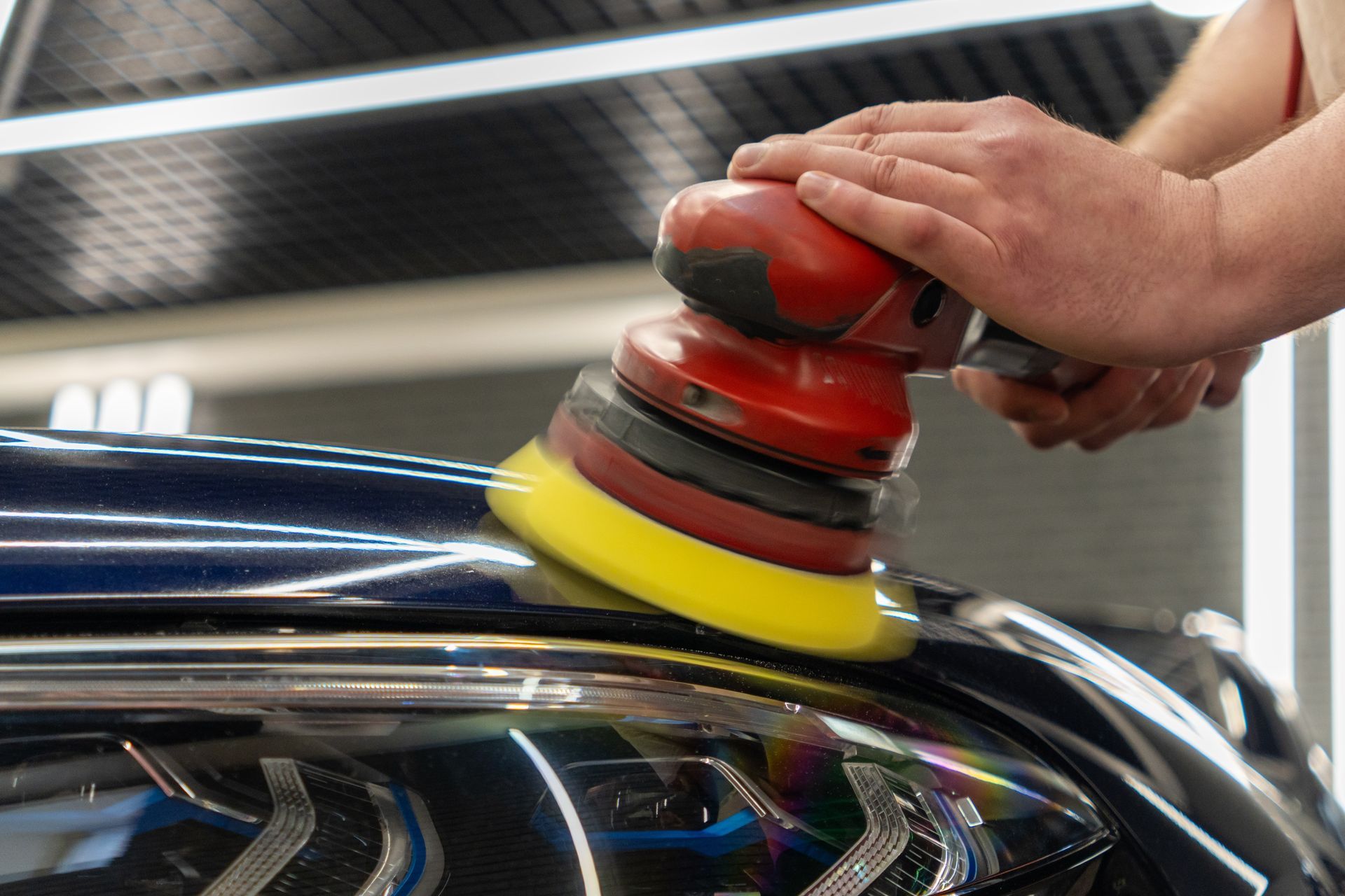 Hands polishing a car with a red-and-black buffer and yellow pad on a glossy hood