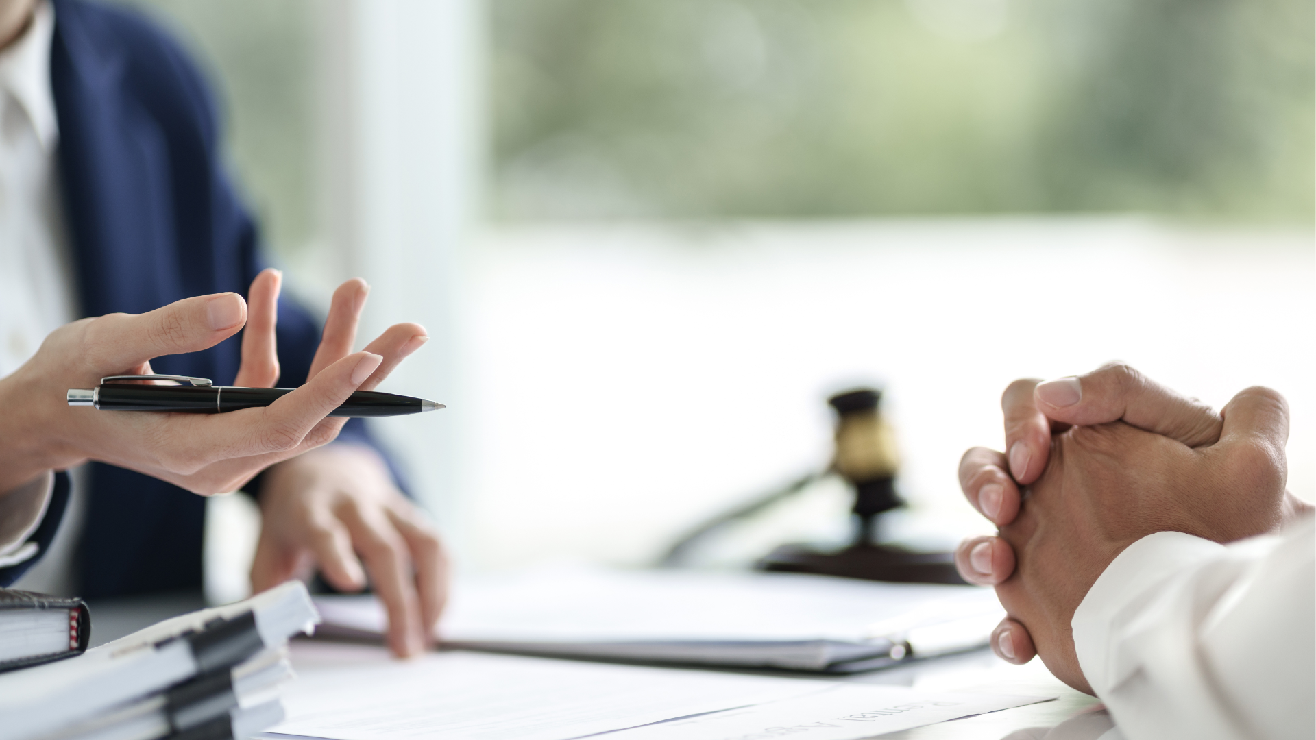 Person gesturing with pen, discussing documents with another; gavel in background.