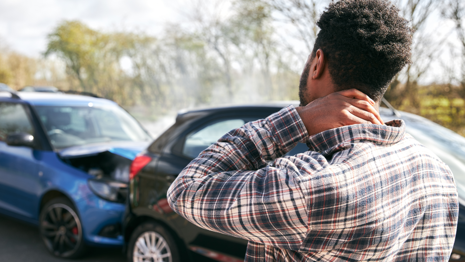 Man holding neck after a car accident, two damaged cars visible.
