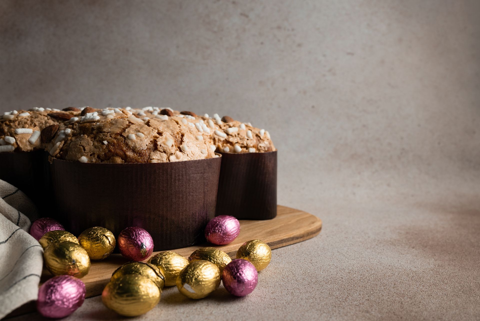 Pane pasquale con noci e uova di cioccolato avvolte nella carta stagnola su un tagliere di legno, su uno sfondo neutro.