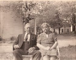 Two people seated outdoors on chairs in a grassy yard with trees and a house behind them.