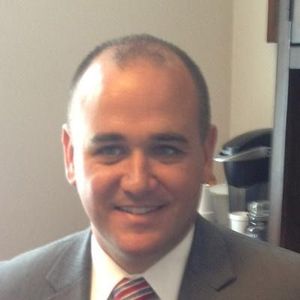 Smiling man in a gray suit and red tie, seated indoors near a doorway